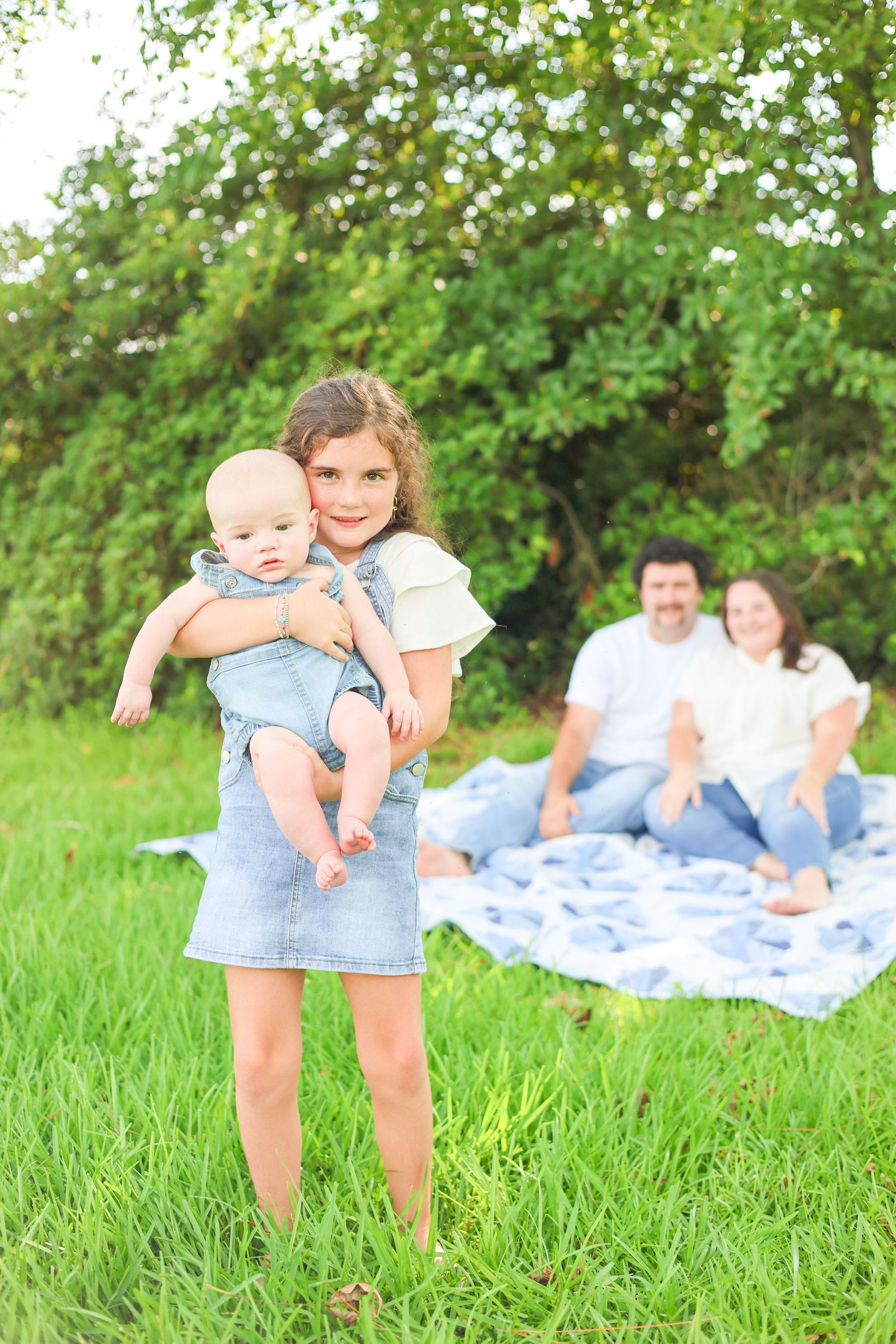 Young girl holding baby sibling outdoors on green grass, parents relaxing on blanket in background.