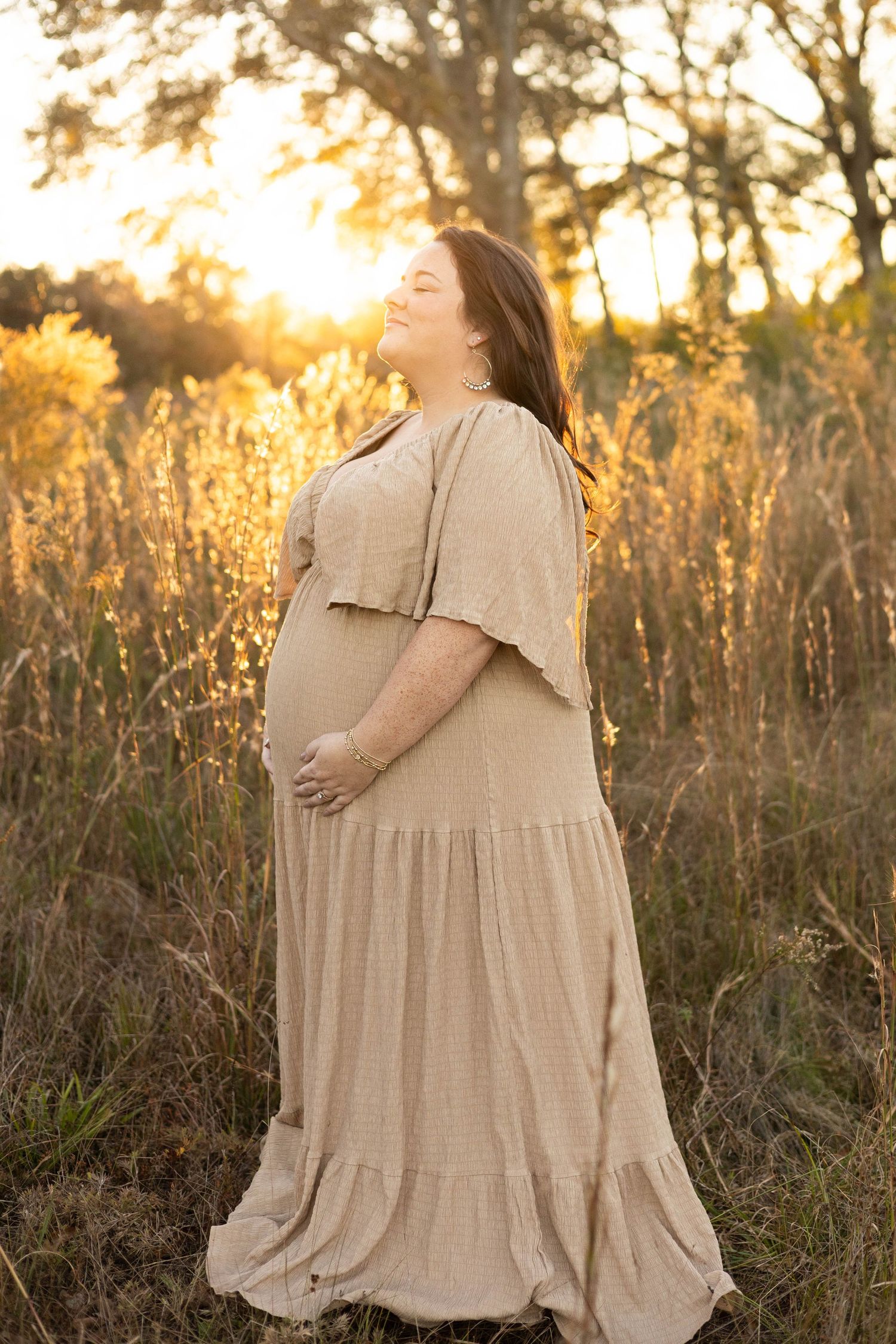 Pregnant woman in beige maxi dress standing in golden field at sunset, smiling and cradling her baby bump.