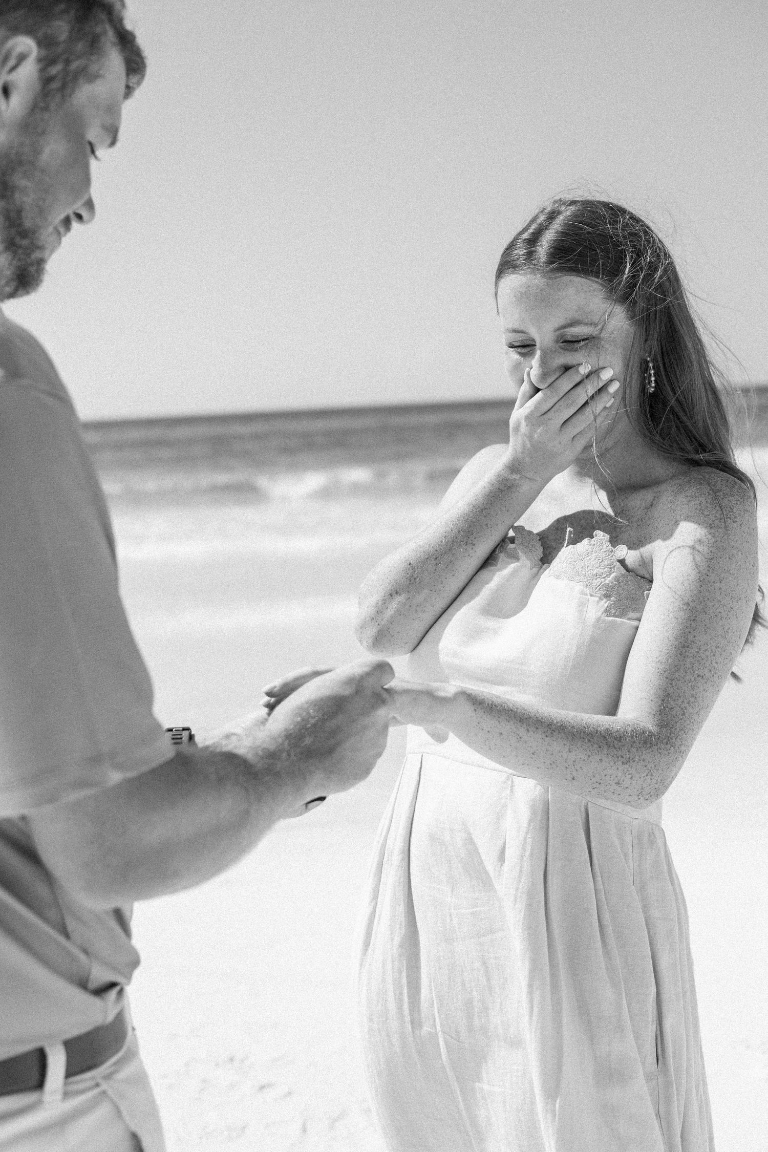 Emotional bride covers her mouth as groom places ring on her finger at beach ceremony.