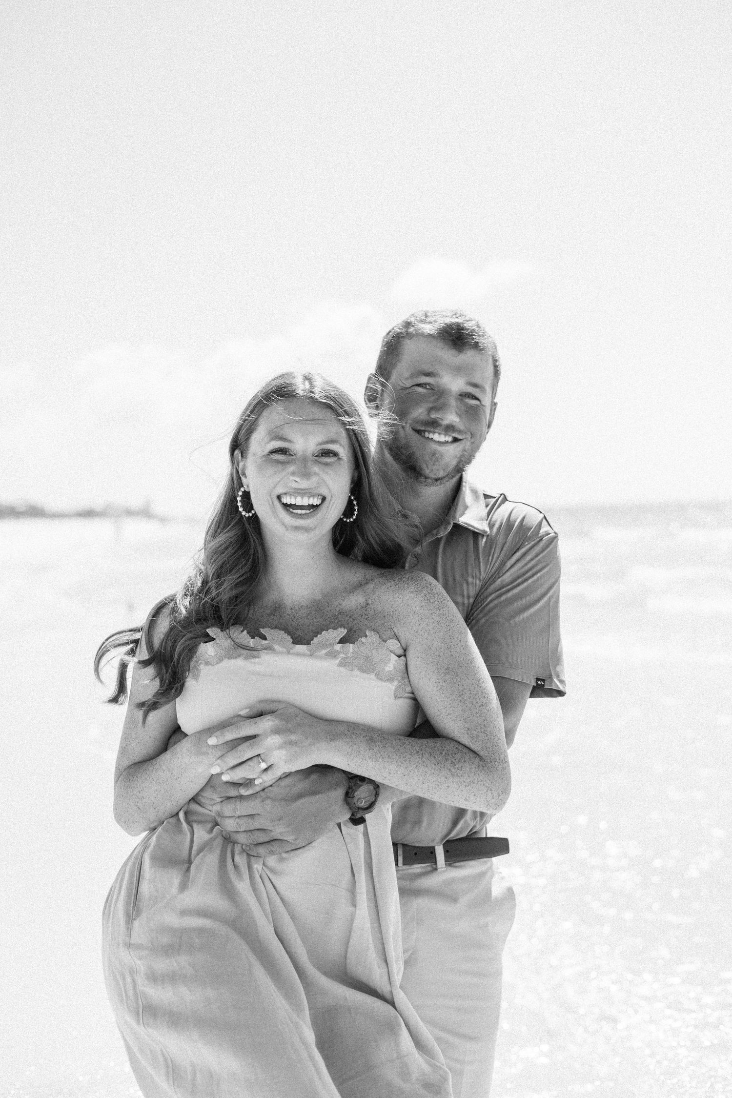Smiling couple embracing on a bright beach, man hugging woman from behind in black and white photo.