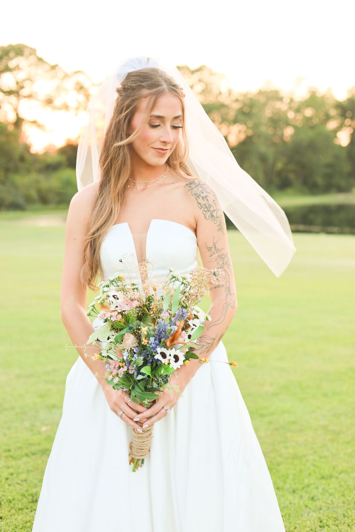 Bride with flowing white dress holds vibrant wildflower bouquet in sunlit field.