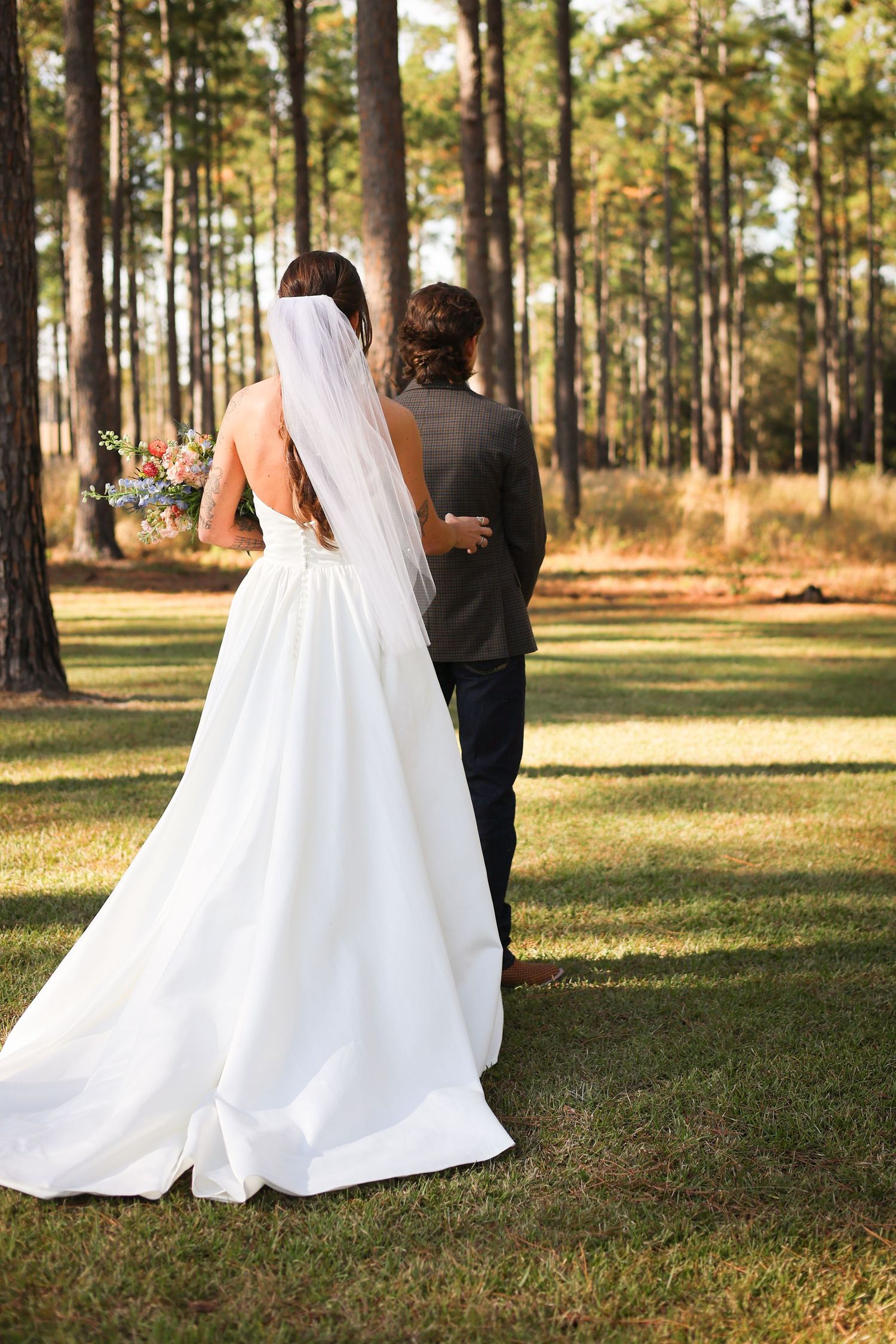 Wedding couple walks through a sunlit pine forest with flowing white dress and veil.