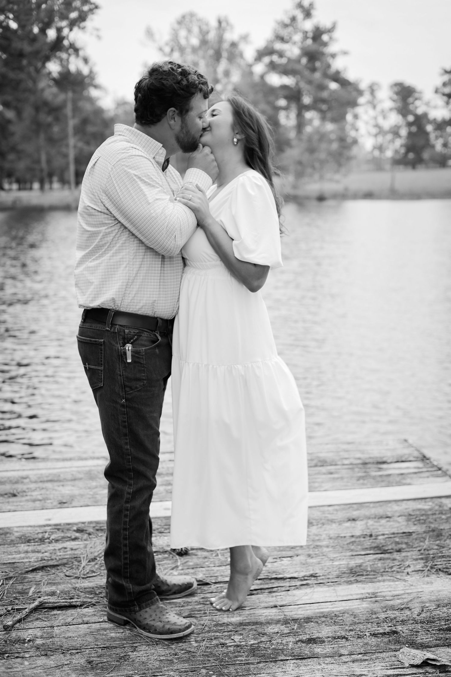 A romantic black and white photo of a couple embracing on a dock by the water.