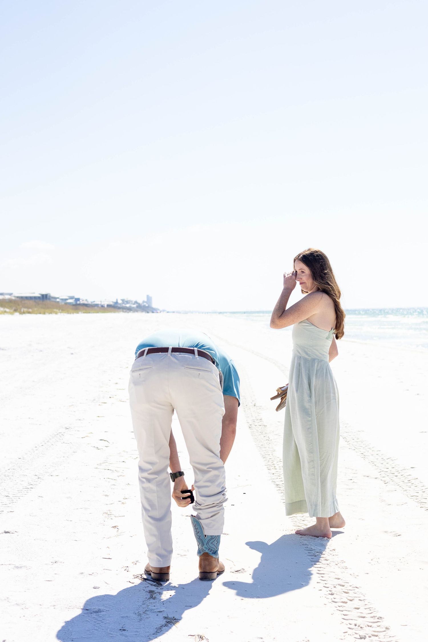Man kneeling on white sand beach while woman in mint dress shields her eyes during a surprise proposal.