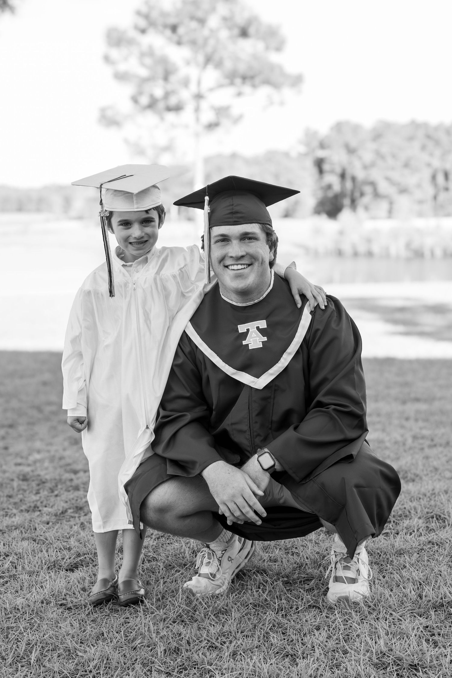 Two graduates in caps and gowns pose together outdoors, one kneeling beside a young child, both smiling.