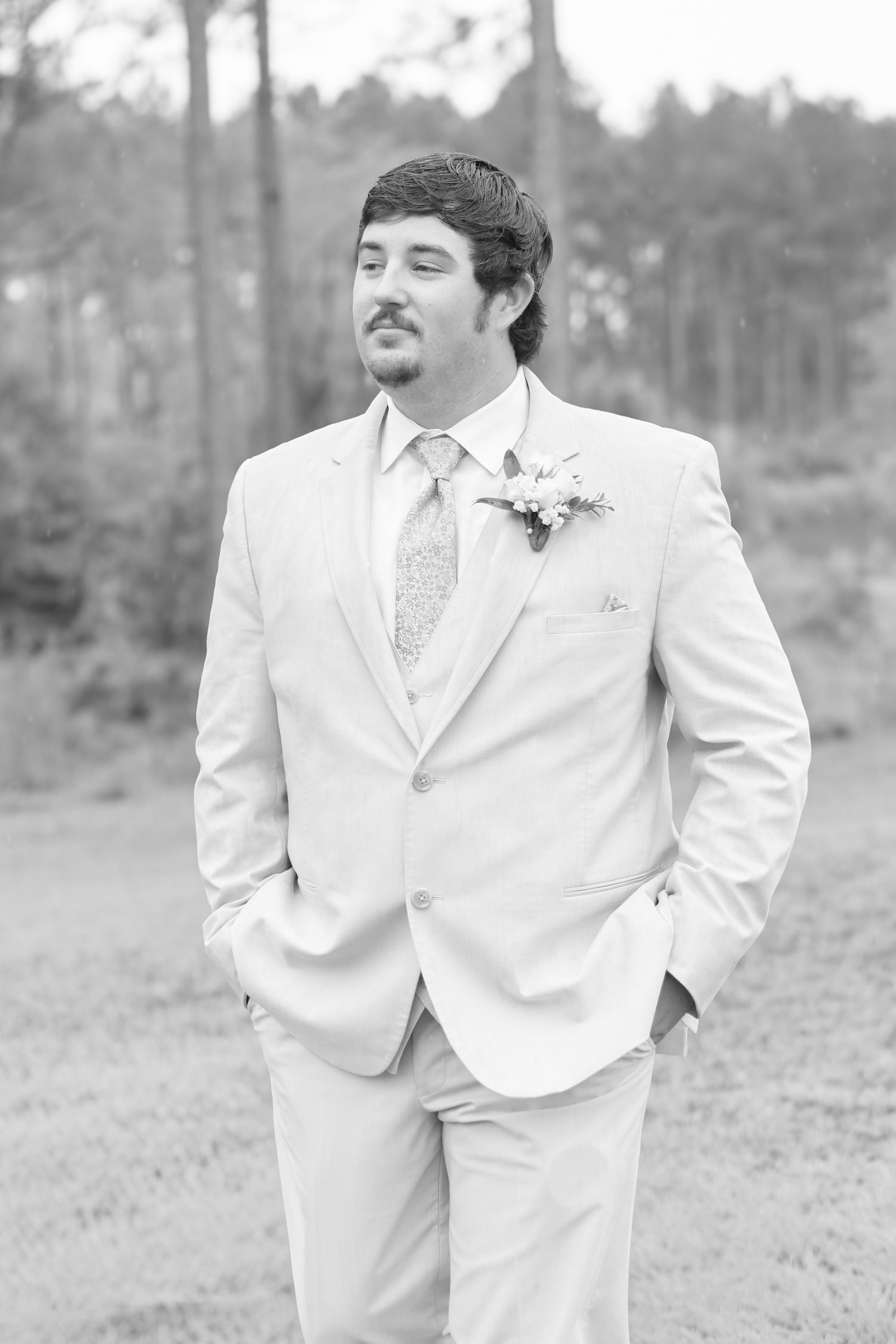 Groom in light suit with boutonniere and tie poses outdoors in black and white wedding photo.
