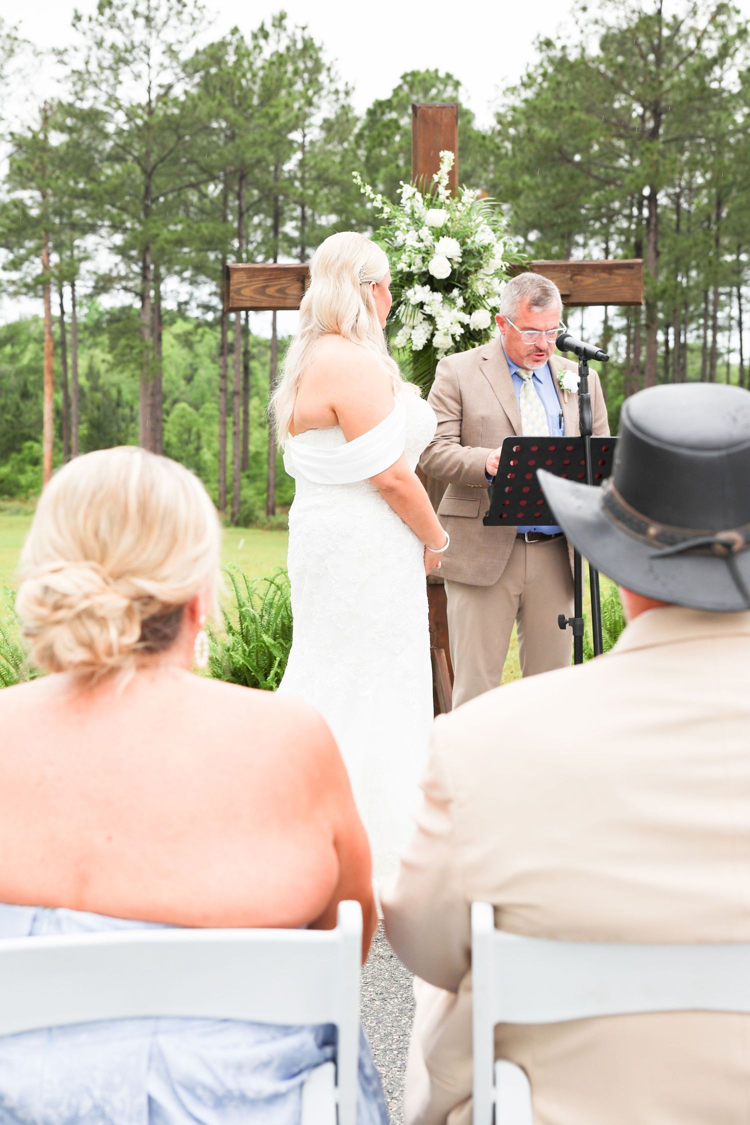 Outdoor wedding ceremony with bride and groom at rustic wooden cross altar, guests seated on white chairs among pine trees.