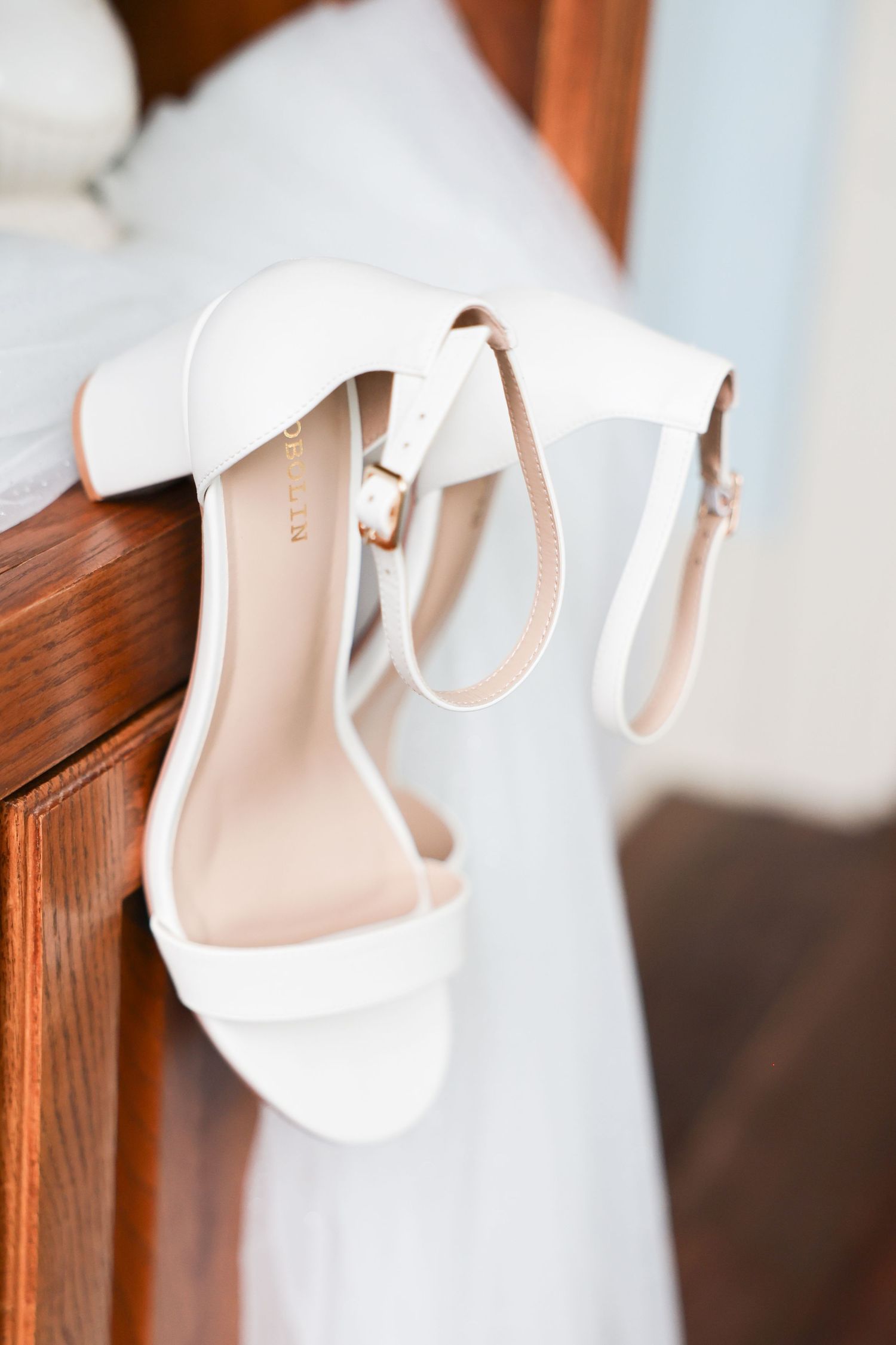 White bridal heeled sandals with ankle straps displayed on a wooden furniture edge with veil.