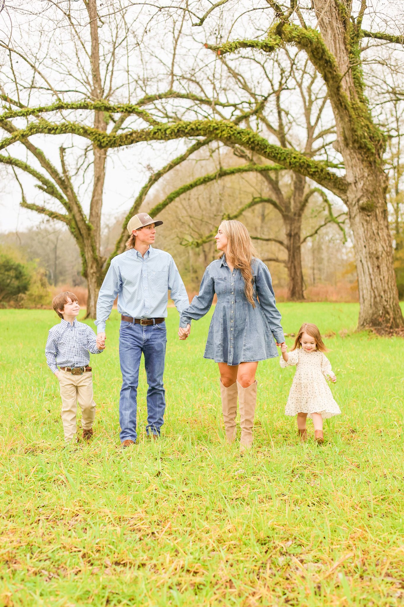 A family of four walks hand in hand through a mossy tree-lined field on an autumn day.