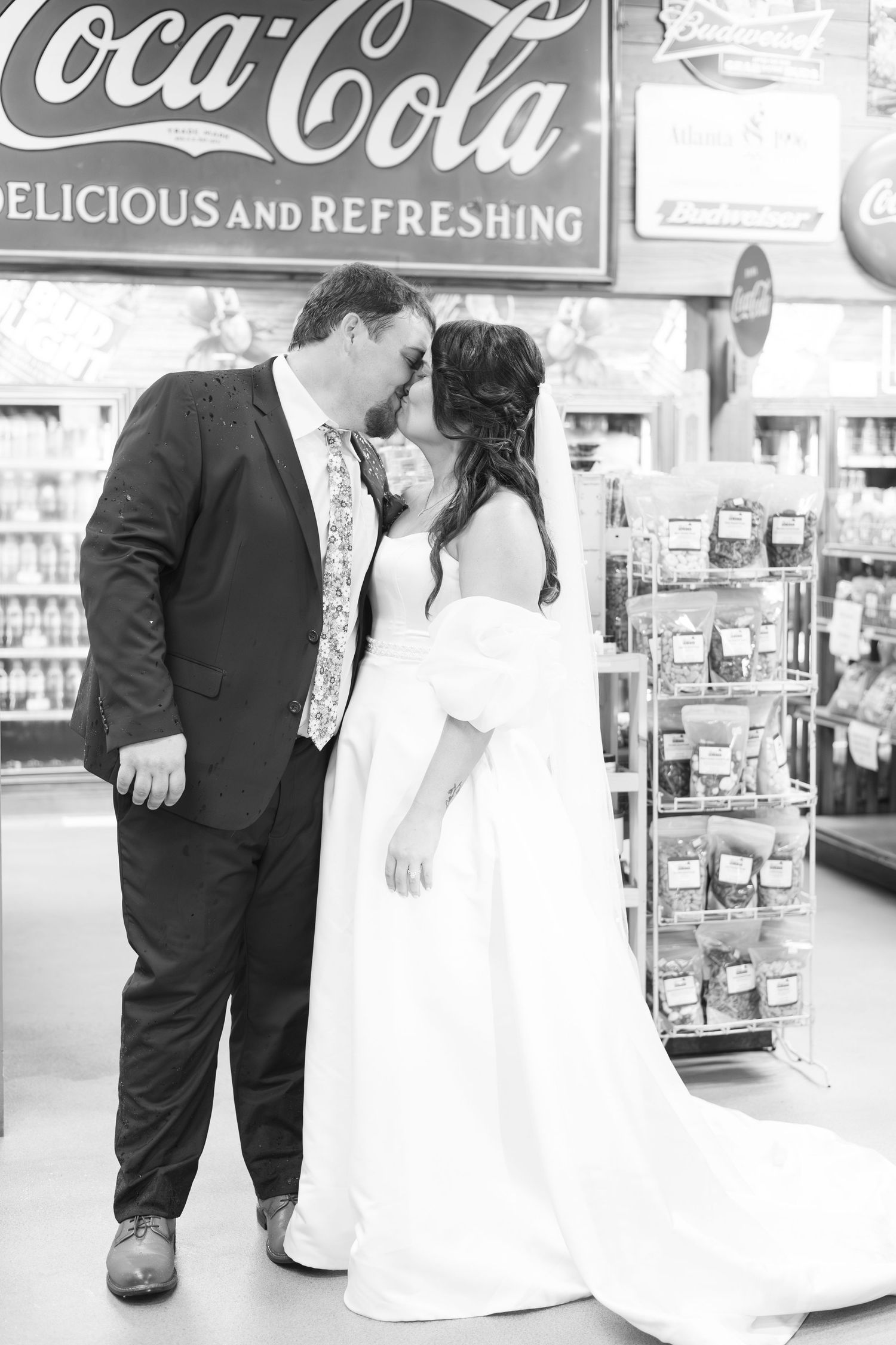 Bride and groom kissing inside a store under a vintage Coca-Cola sign, black and white photo.