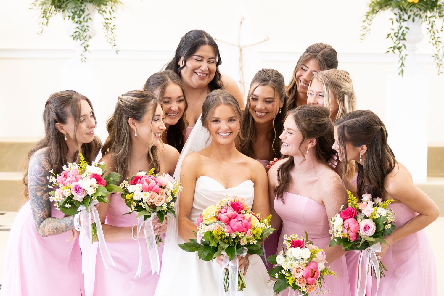 Bride in white strapless gown surrounded by bridesmaids in pink dresses holding colorful floral bouquets.
