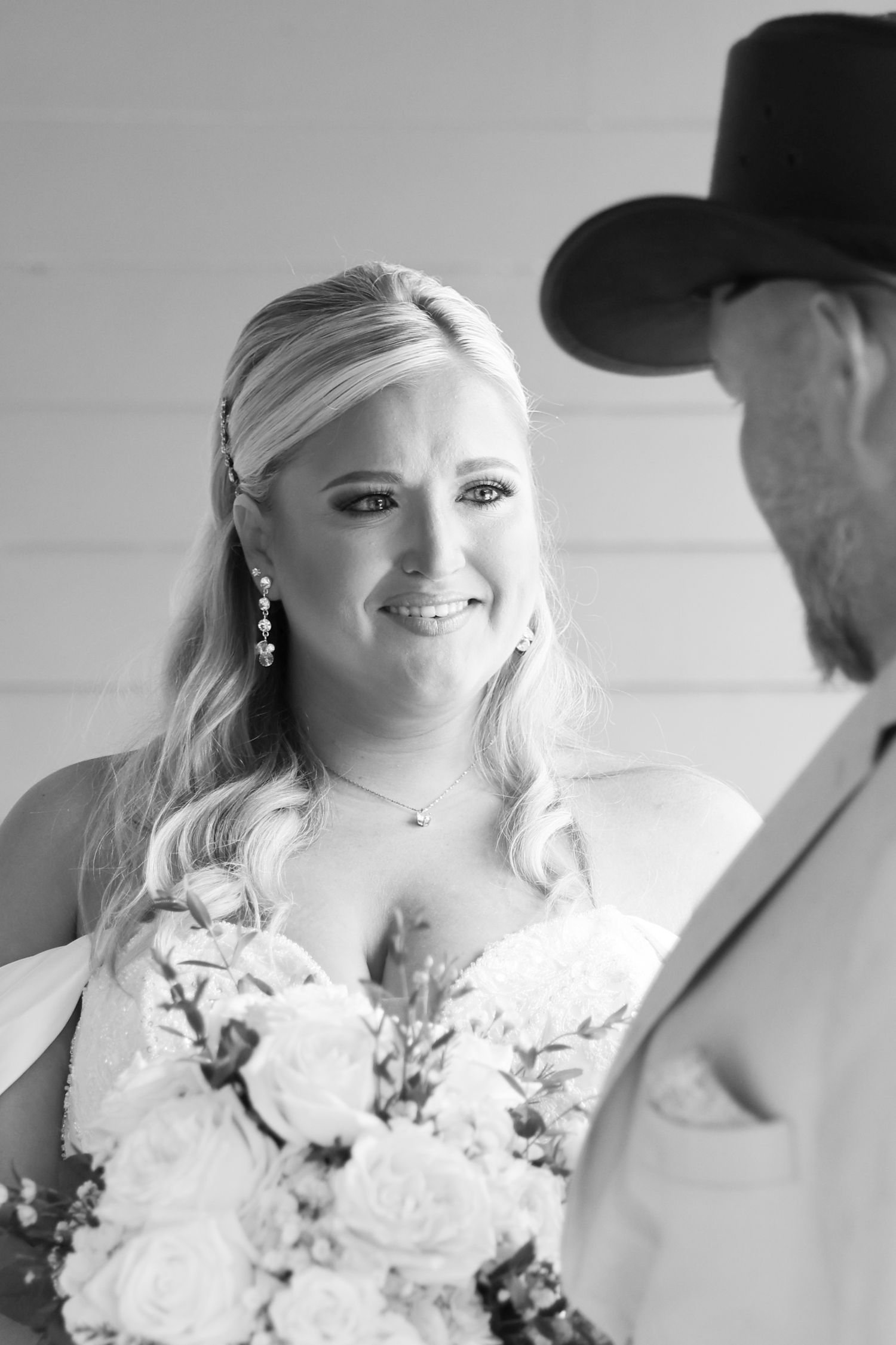 Black and white photo of a smiling bride holding a bouquet, facing a man in a cowboy hat at a wedding ceremony.