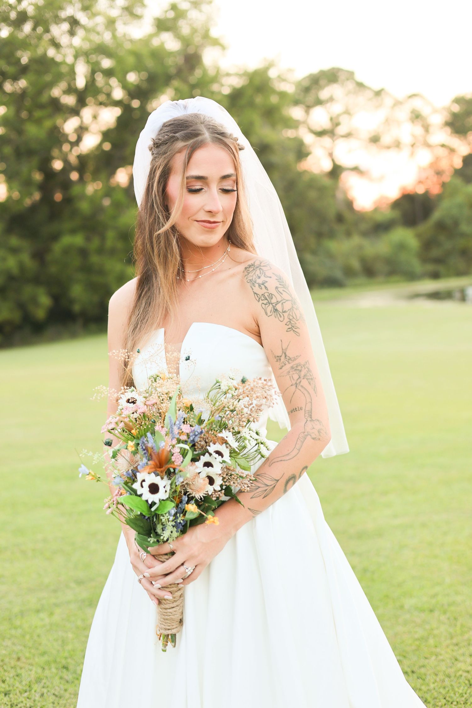 Tattooed bride in strapless white gown and veil holds wildflower bouquet outdoors at golden hour.