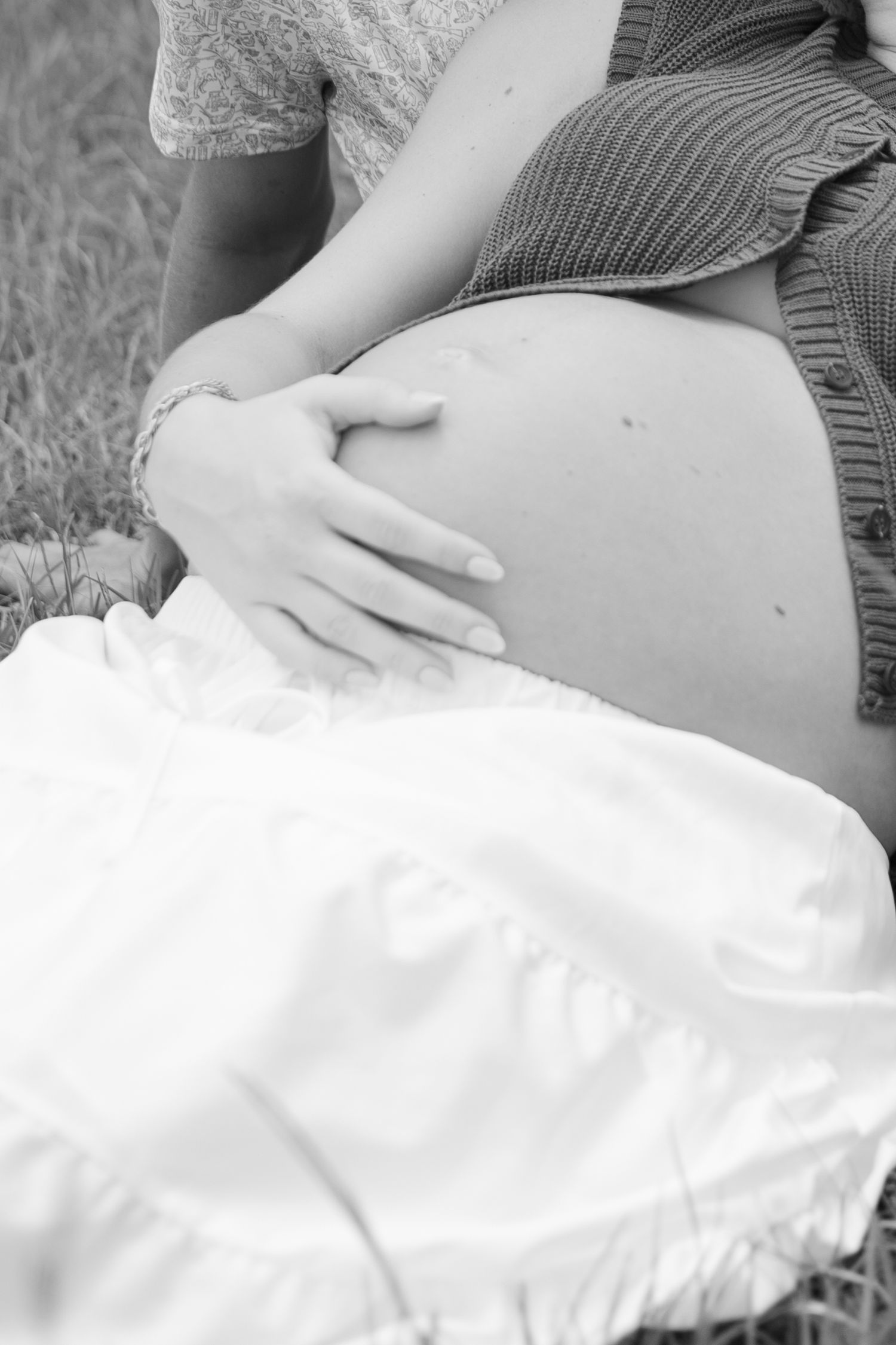 Black and white photo of a pregnant woman cradling her bare baby bump while sitting outdoors in a field.