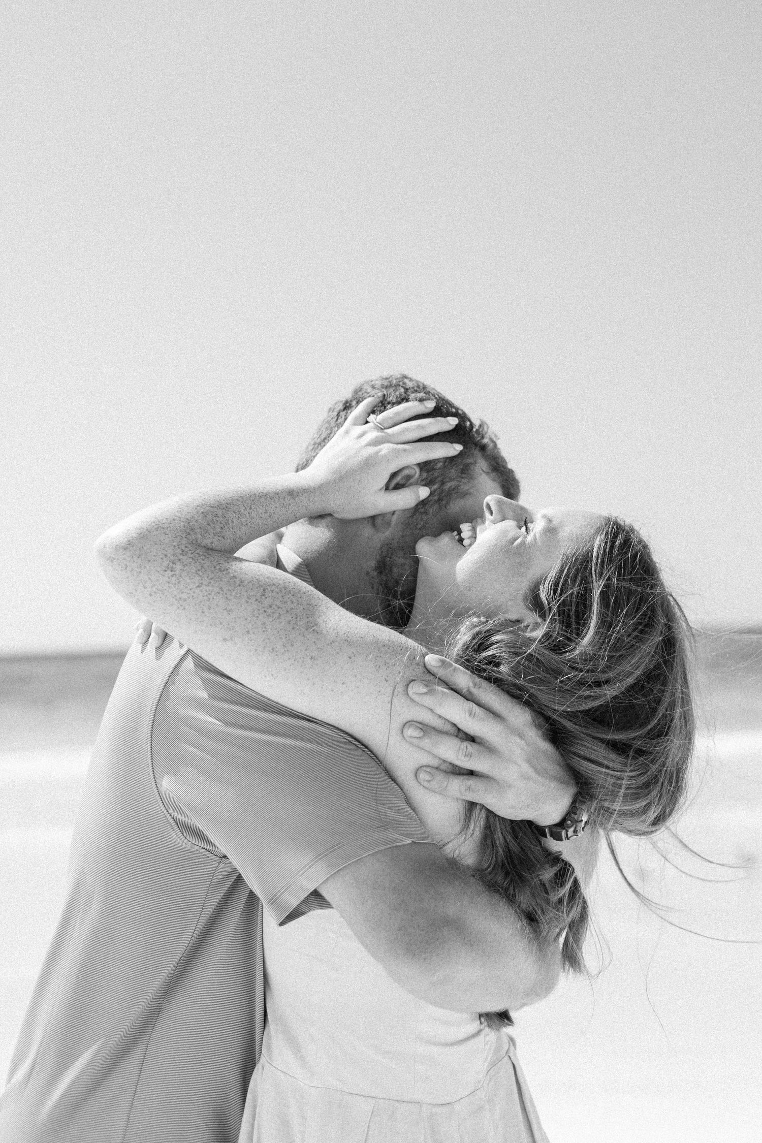 Black and white romantic photo of couple kissing and embracing on a breezy beach.