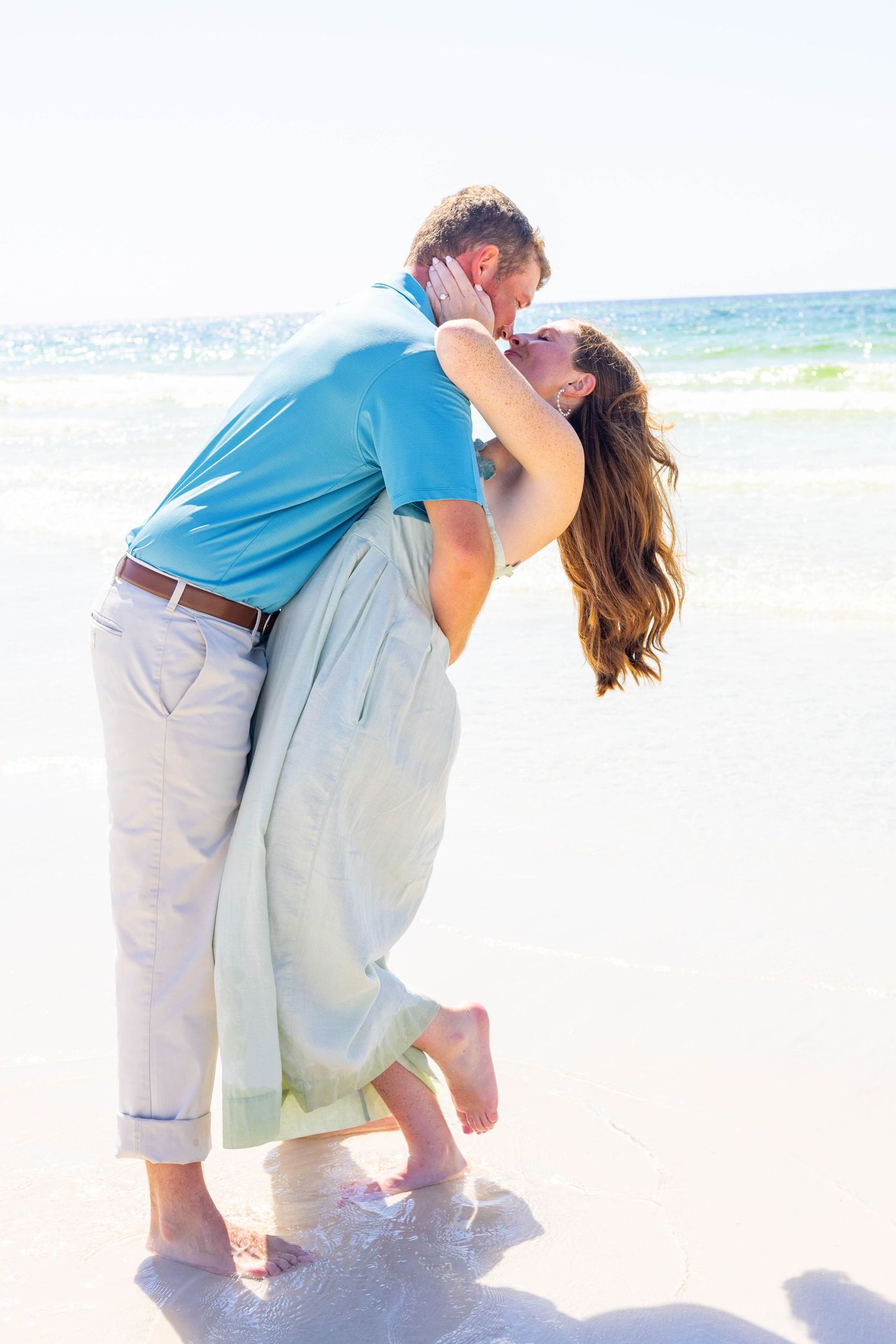 Romantic couple sharing a kiss on a sunny beach, woman in flowing dress and man in blue polo by the ocean waves.