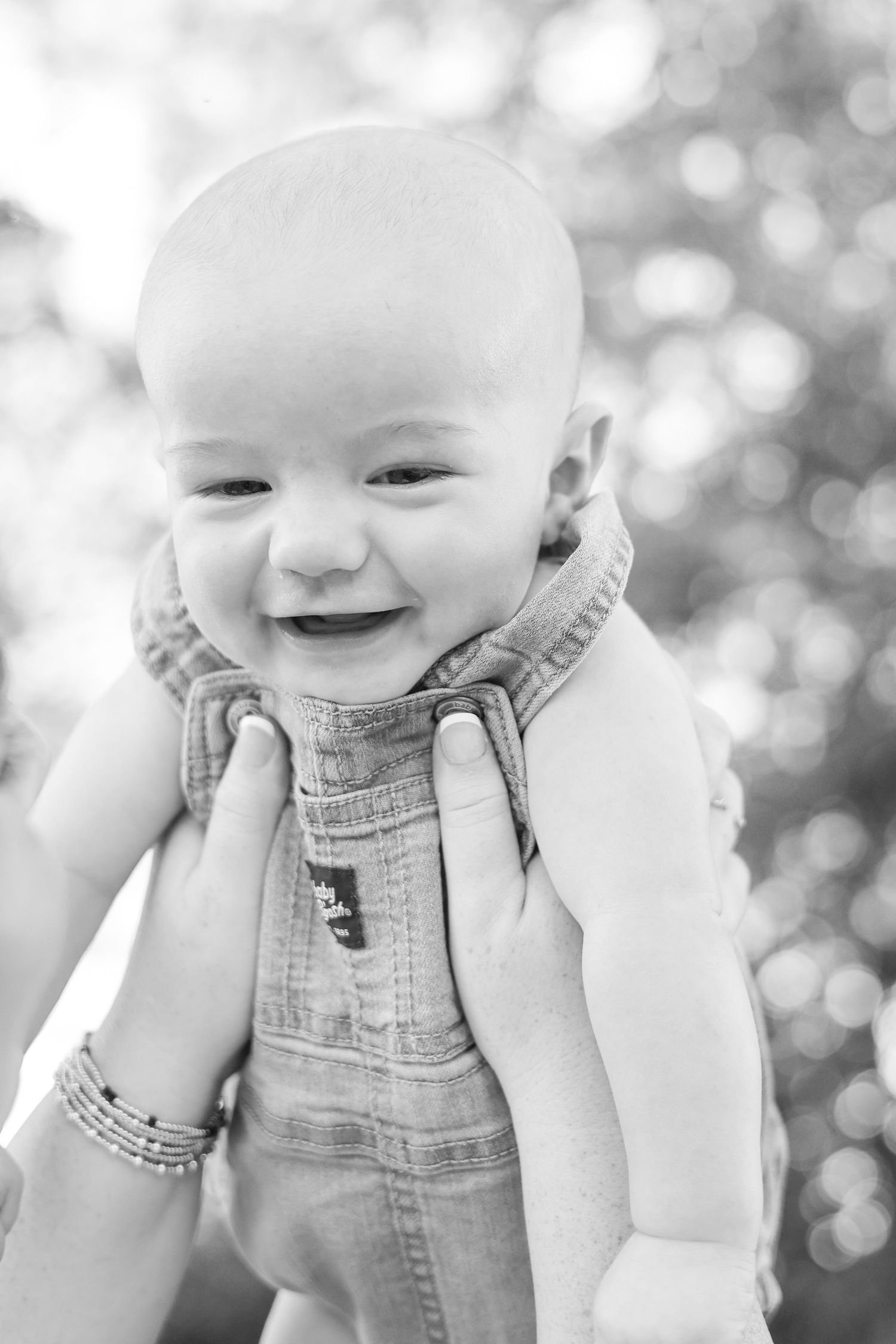 Black and white photo of a smiling bald baby in denim overalls being held up by adult hands outdoors.