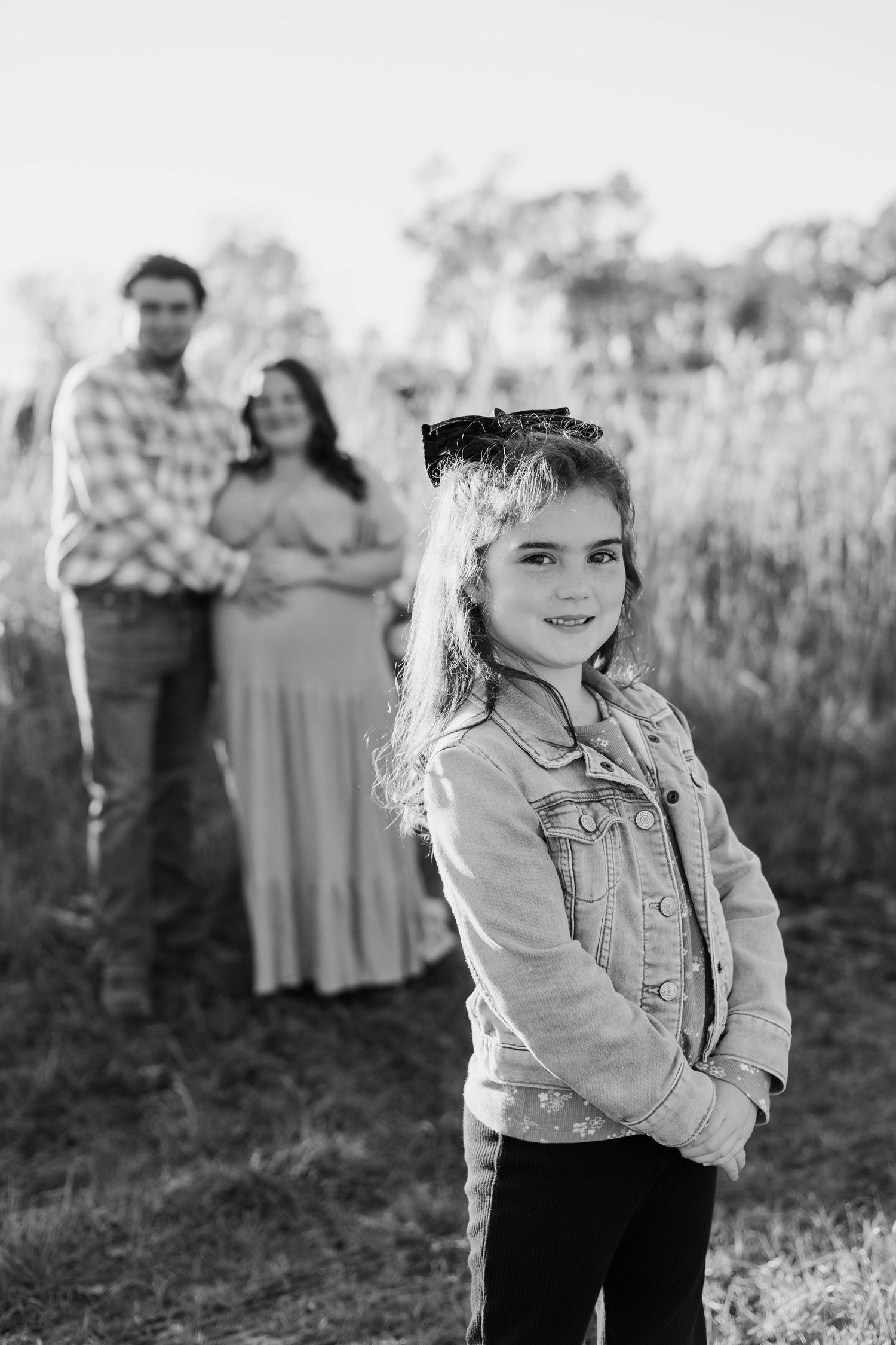 Black and white family maternity photo with young girl smiling in foreground, parents embracing in blurred background outdoors.