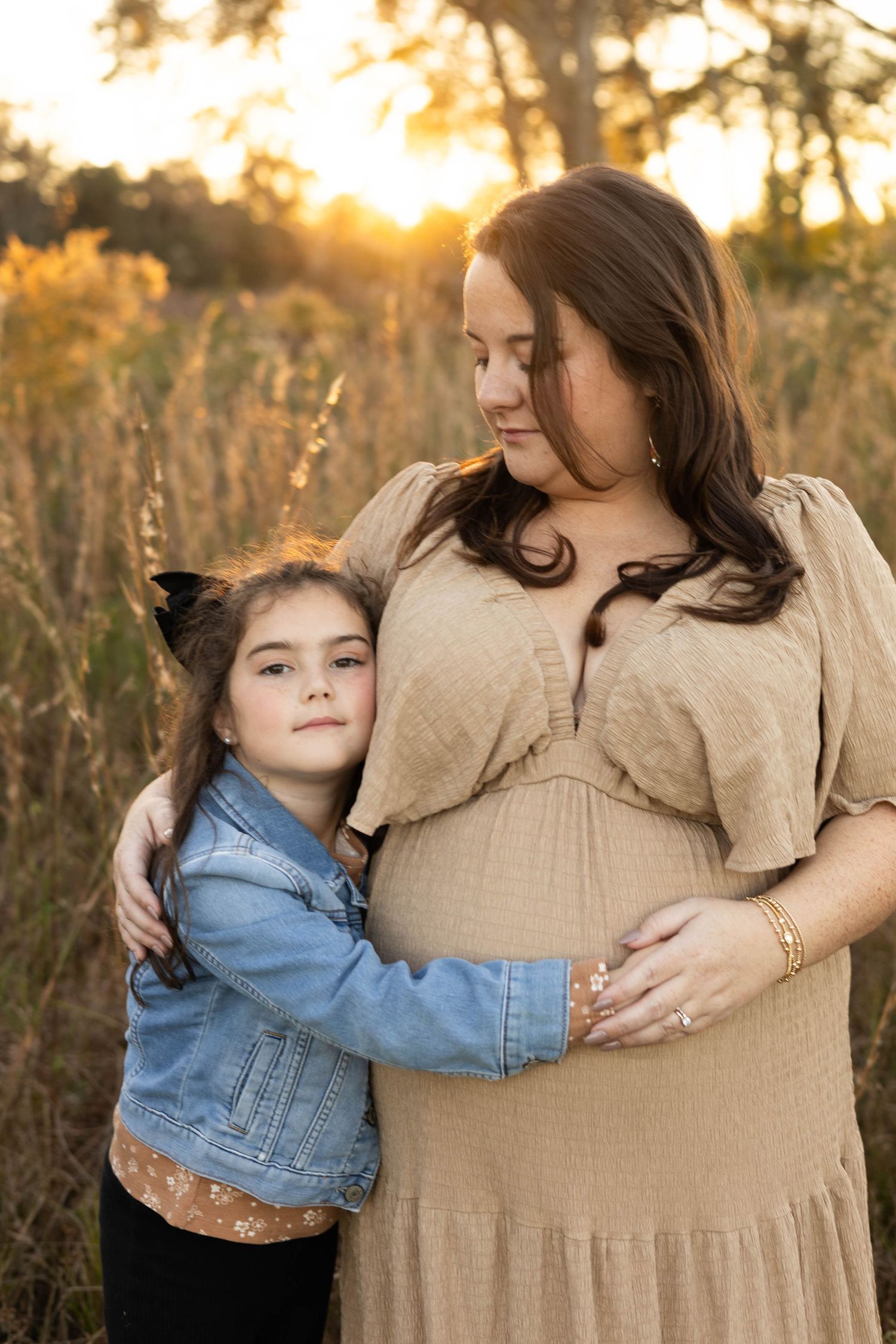 A pregnant woman in a tan dress embraces her young daughter in a golden sunset field.