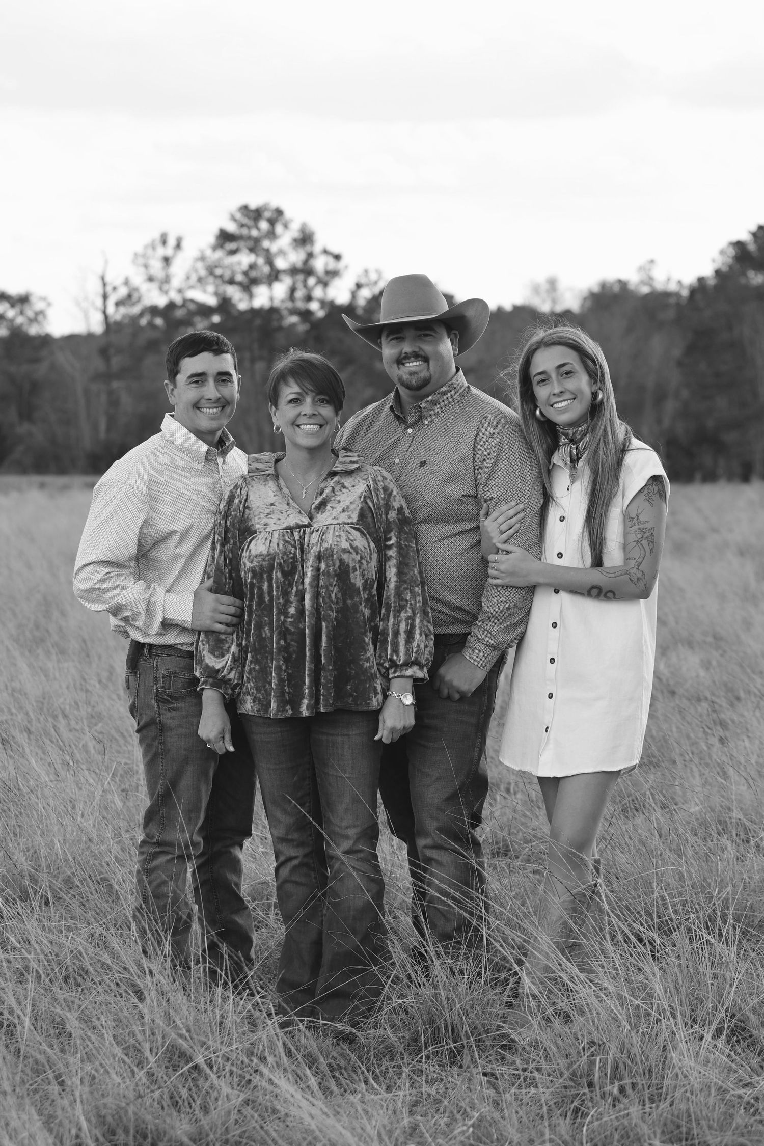 Black and white family portrait of four people smiling in a grassy field, man wearing cowboy hat.