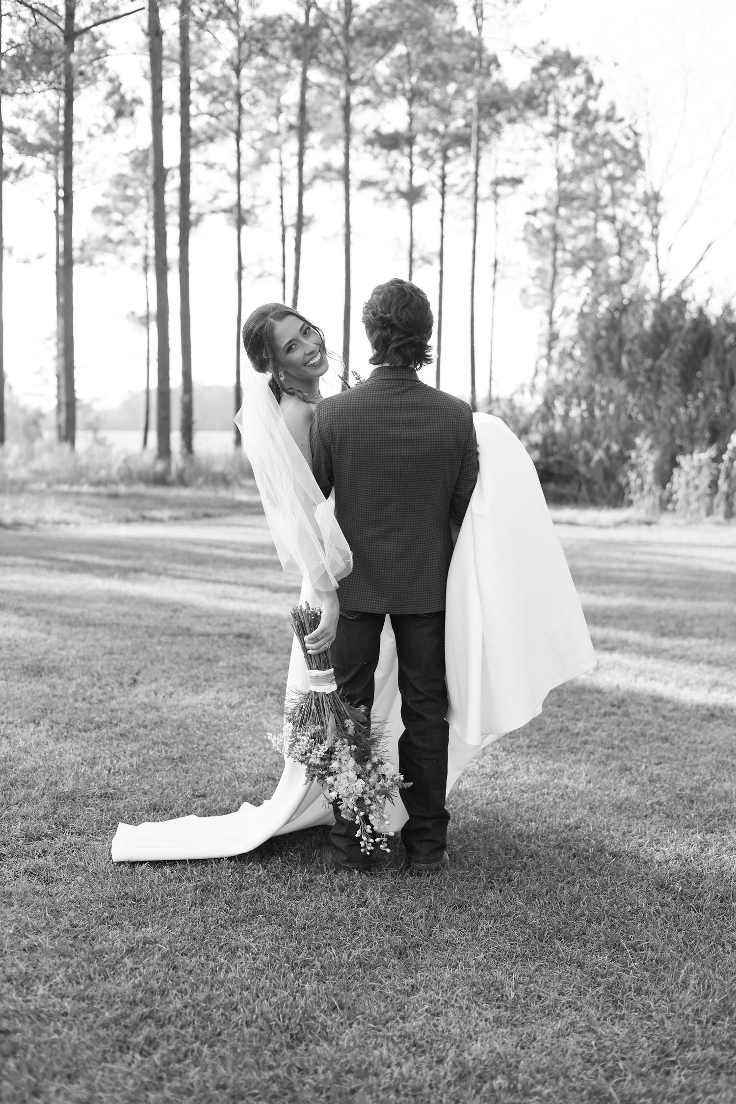 Black and white photo of a smiling bride in a veil holding a bouquet, standing with a groom in a suit outdoors.