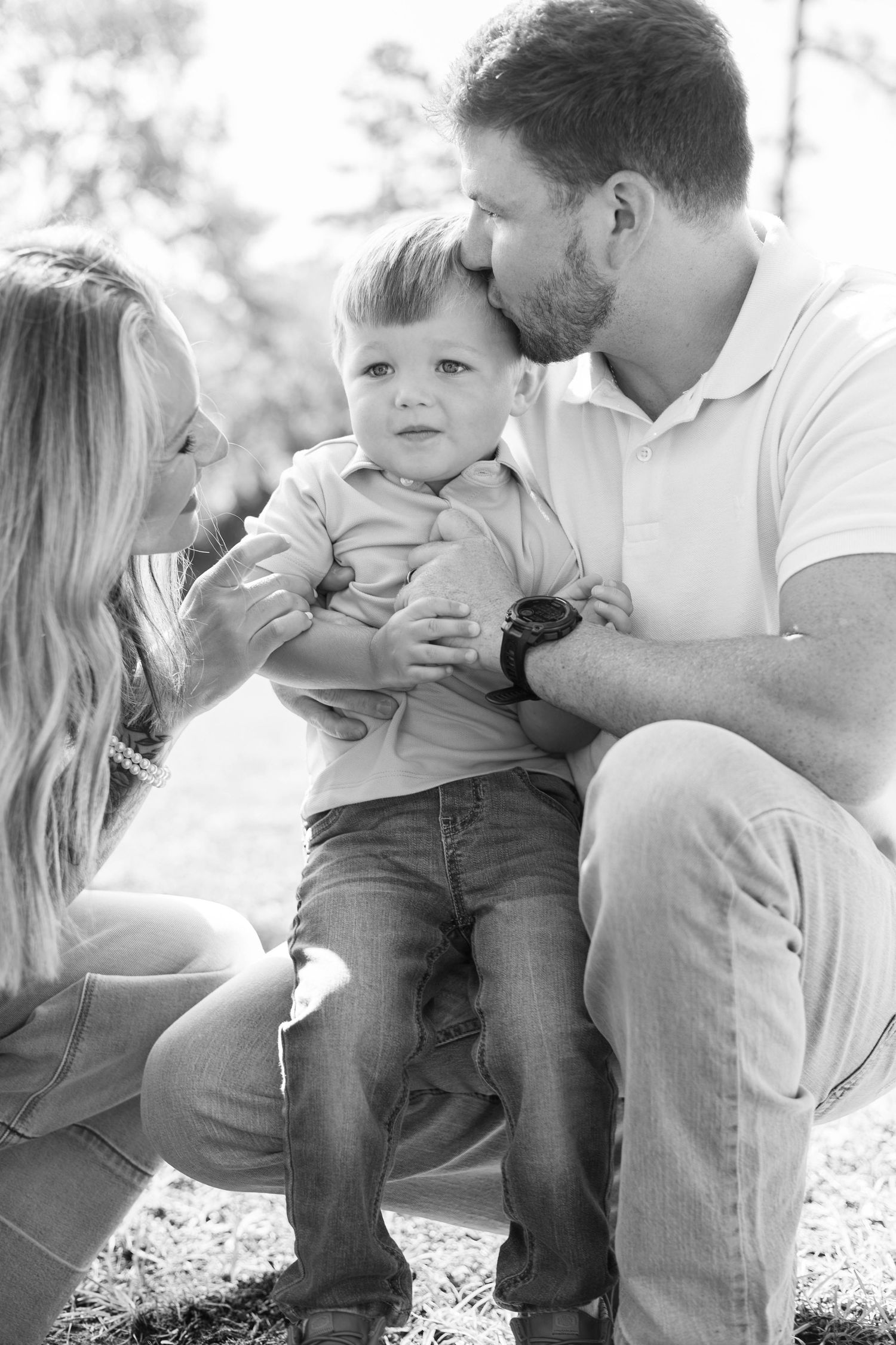 Father kisses toddler son on head while mother smiles in black and white family portrait.