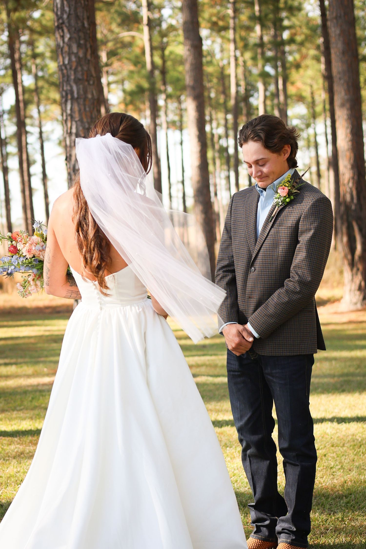 Bride in flowing white gown approaches groom for first look in wooded setting.