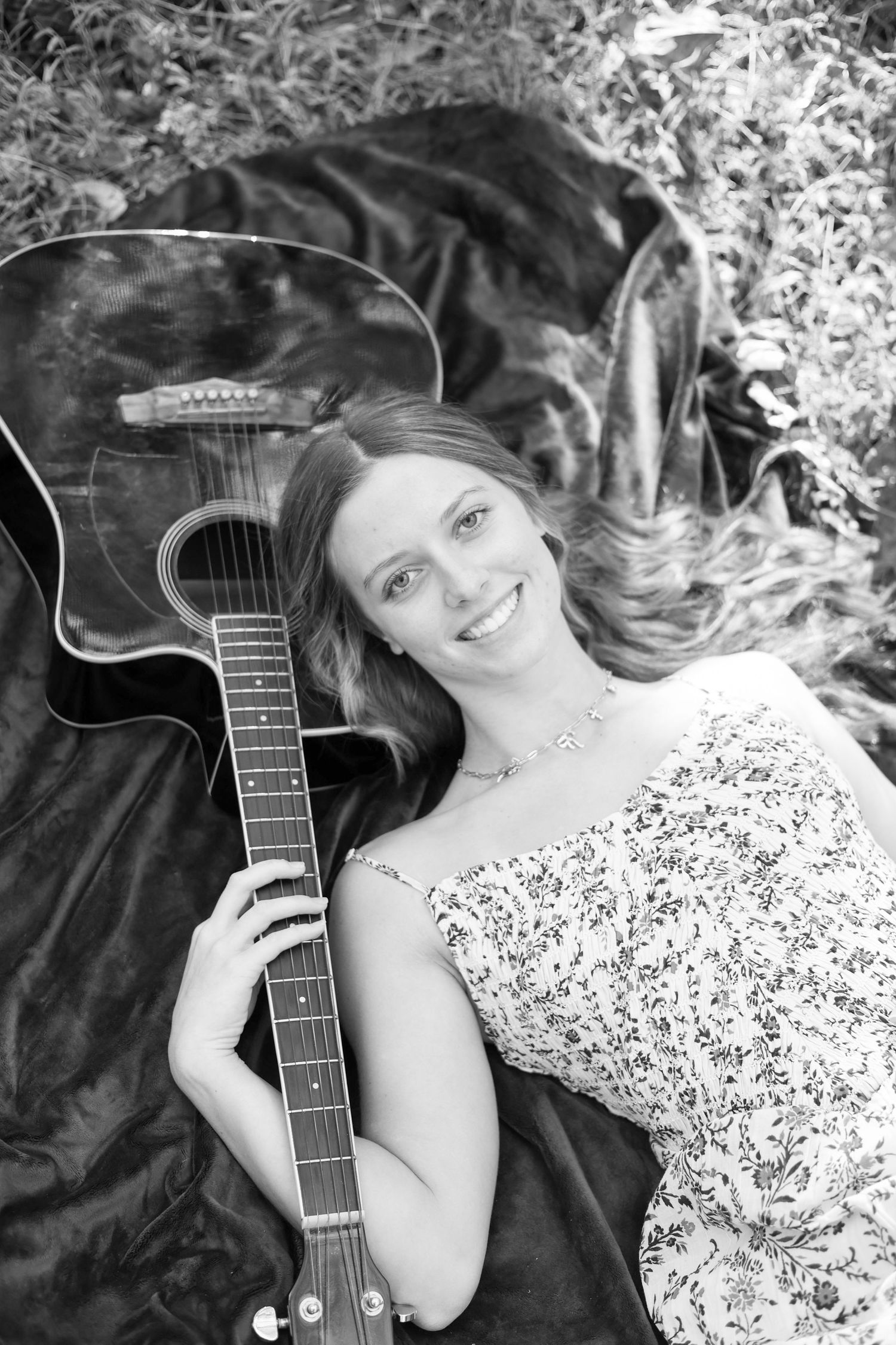 Black and white portrait of person lying down with acoustic guitar in floral dress.