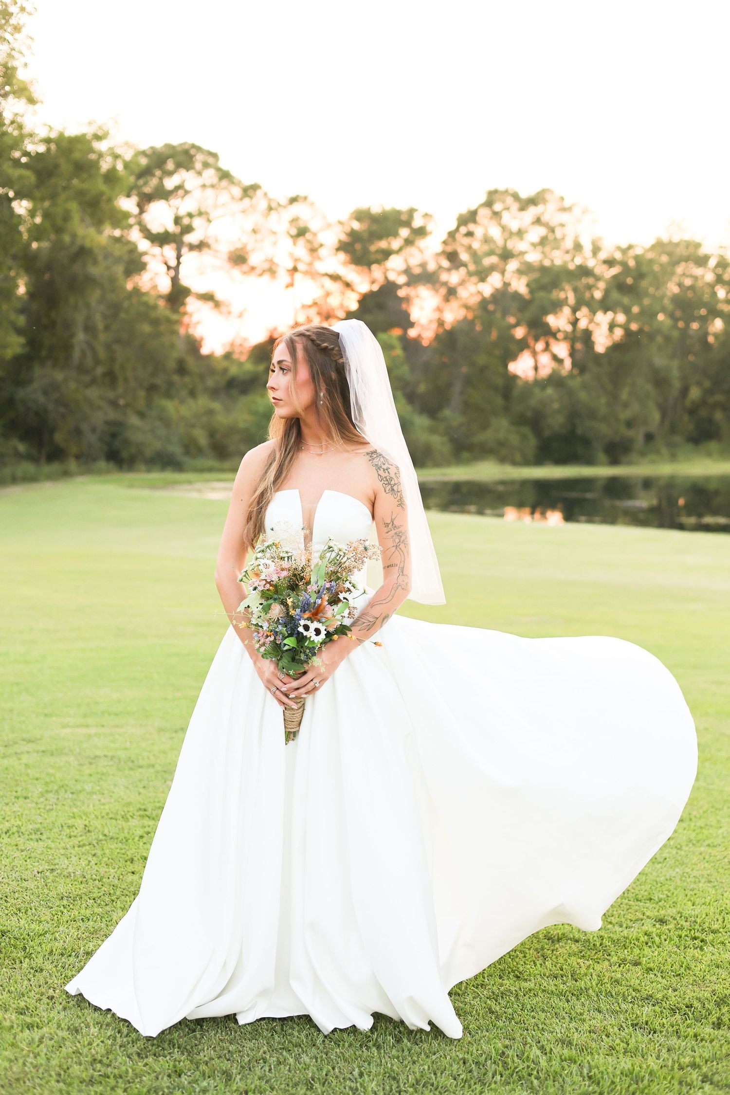 Wedding dress flowing in breeze as bride poses with bouquet in grassy field at sunset.