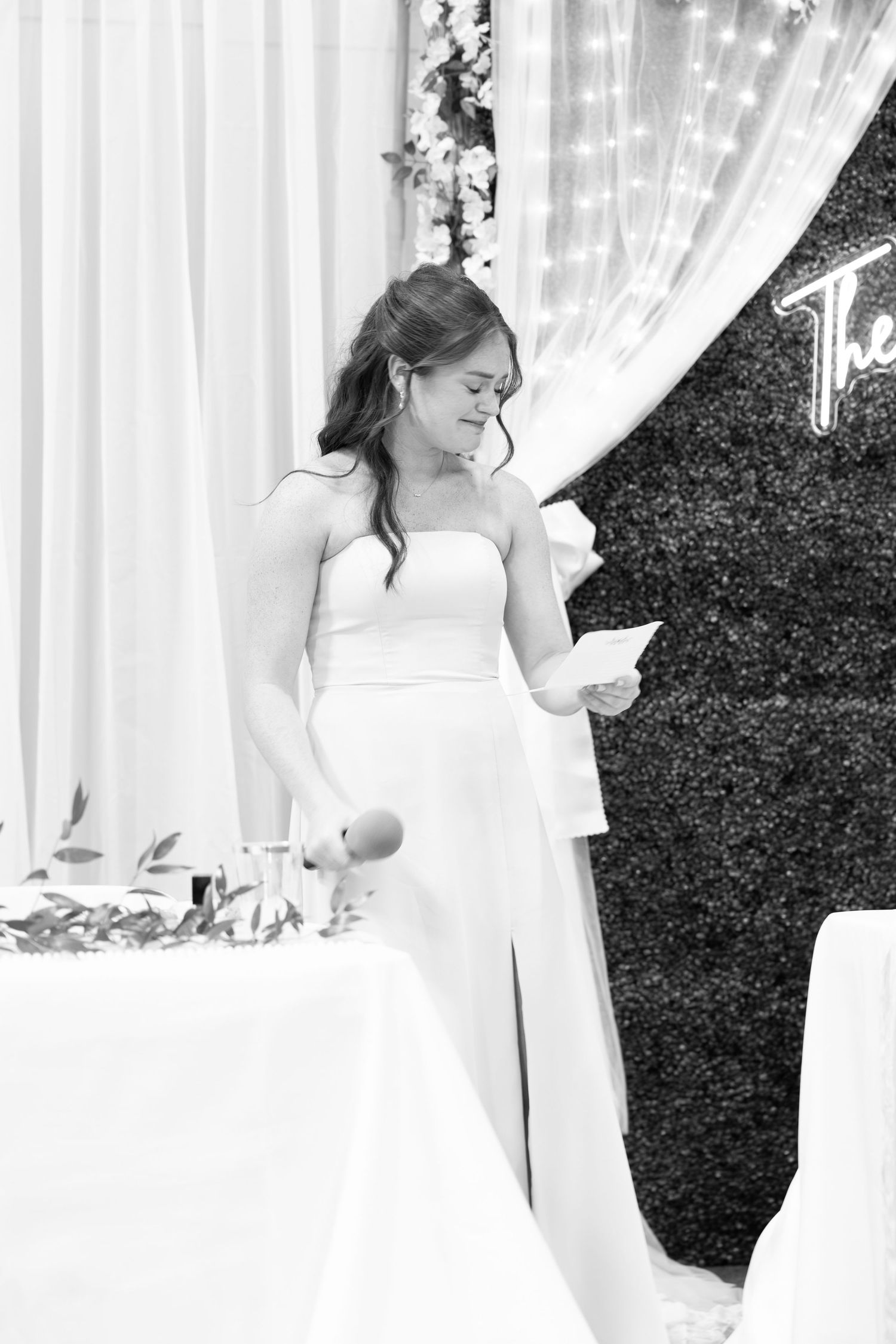 Elegant bride in a strapless white gown reads her vows at a decorated wedding reception venue.