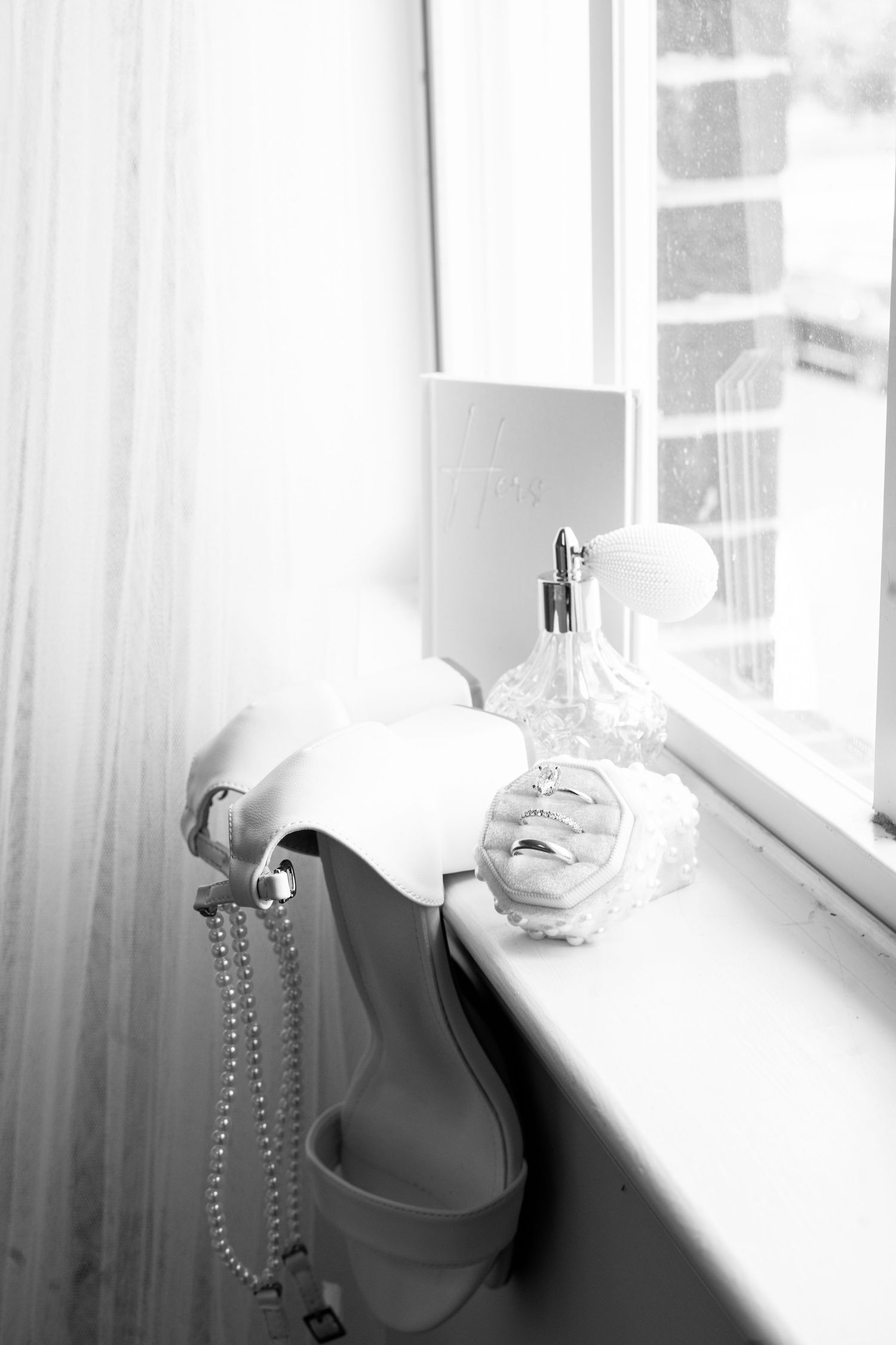 Black and white wedding flat lay with bridal heels, pearl necklace, rings, and perfume bottle on a windowsill.