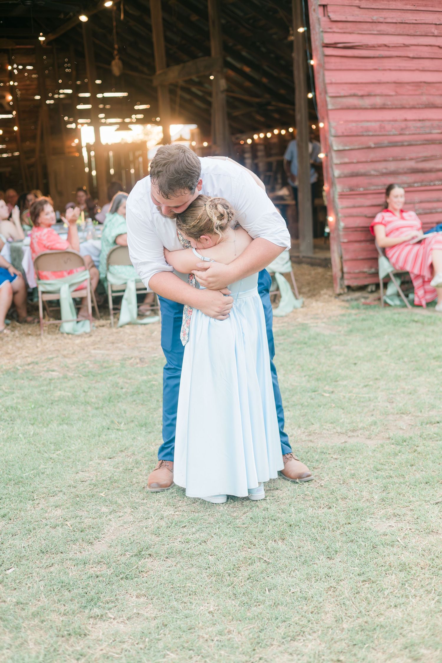 Groom hugging flower girl in light blue dress at outdoor barn wedding reception at sunset.