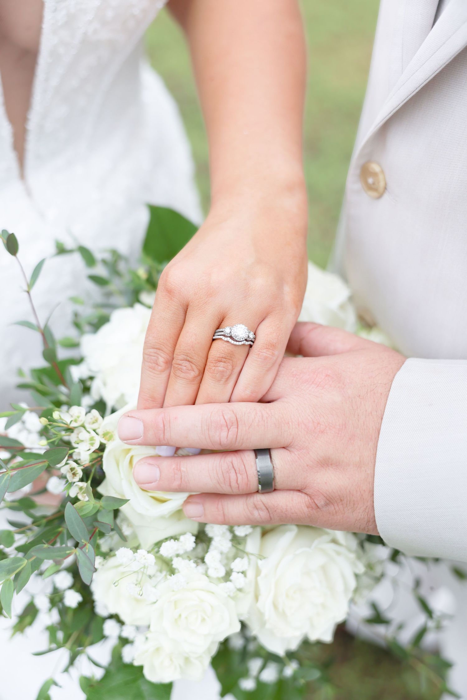 Bride and groom displaying wedding rings over a white rose and greenery bridal bouquet.