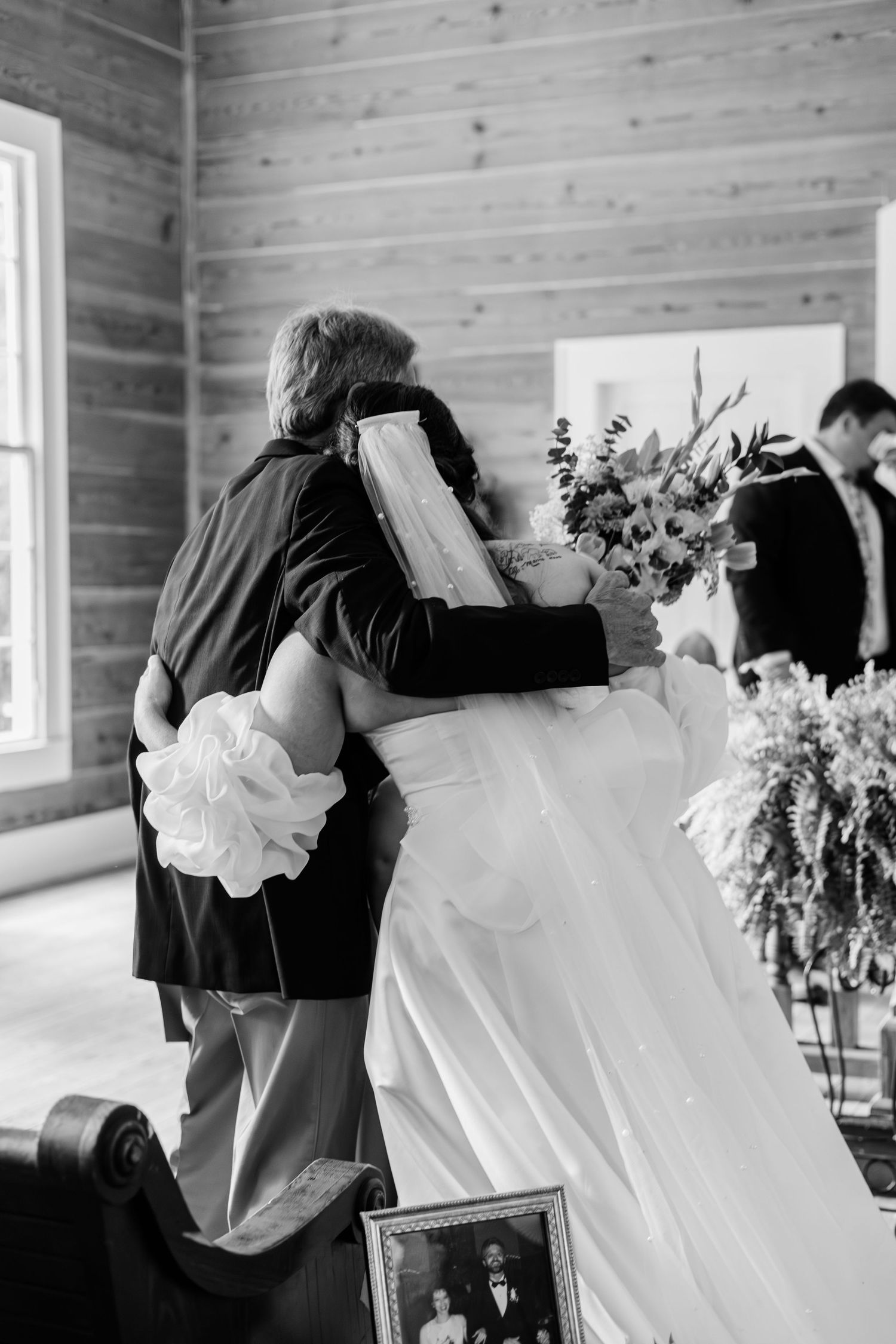 Black and white photo of a bride embracing a family member at a rustic wooden chapel wedding ceremony.