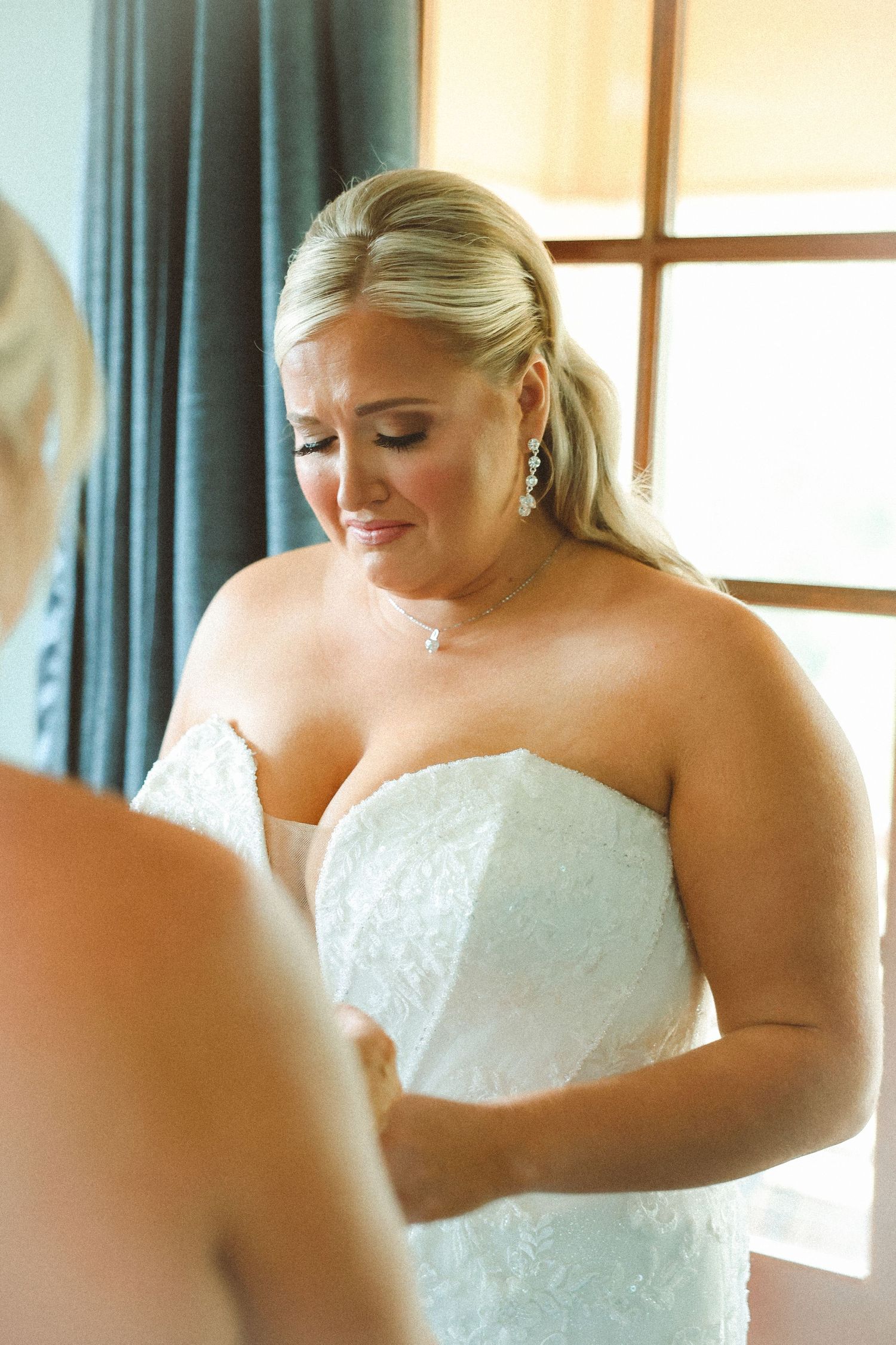 Emotional bride in strapless lace wedding gown being helped to dress before her wedding ceremony.