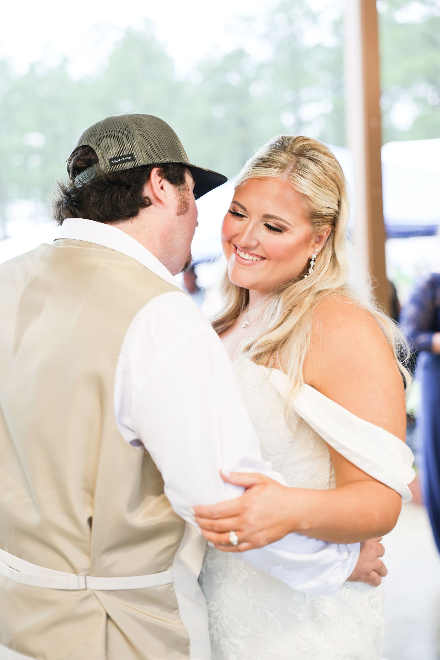 Smiling bride in off-shoulder lace gown shares first dance with groom wearing tan vest and trucker hat.