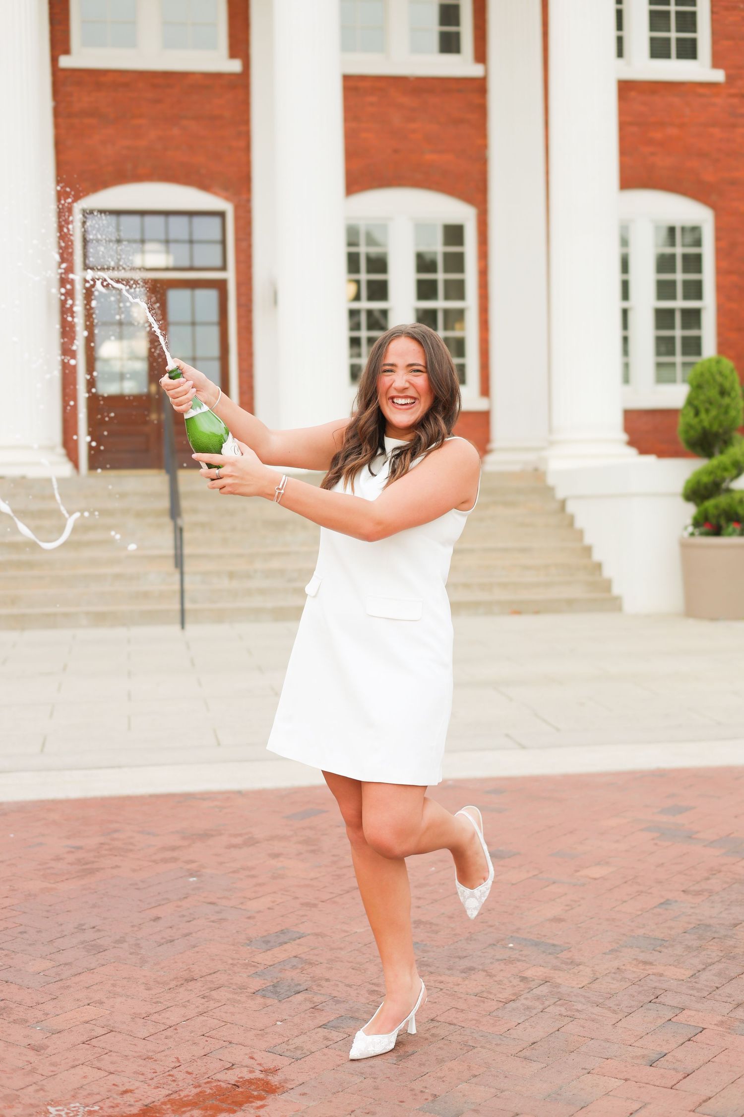 Person in white dress dancing joyfully with champagne outside a collegiate building.