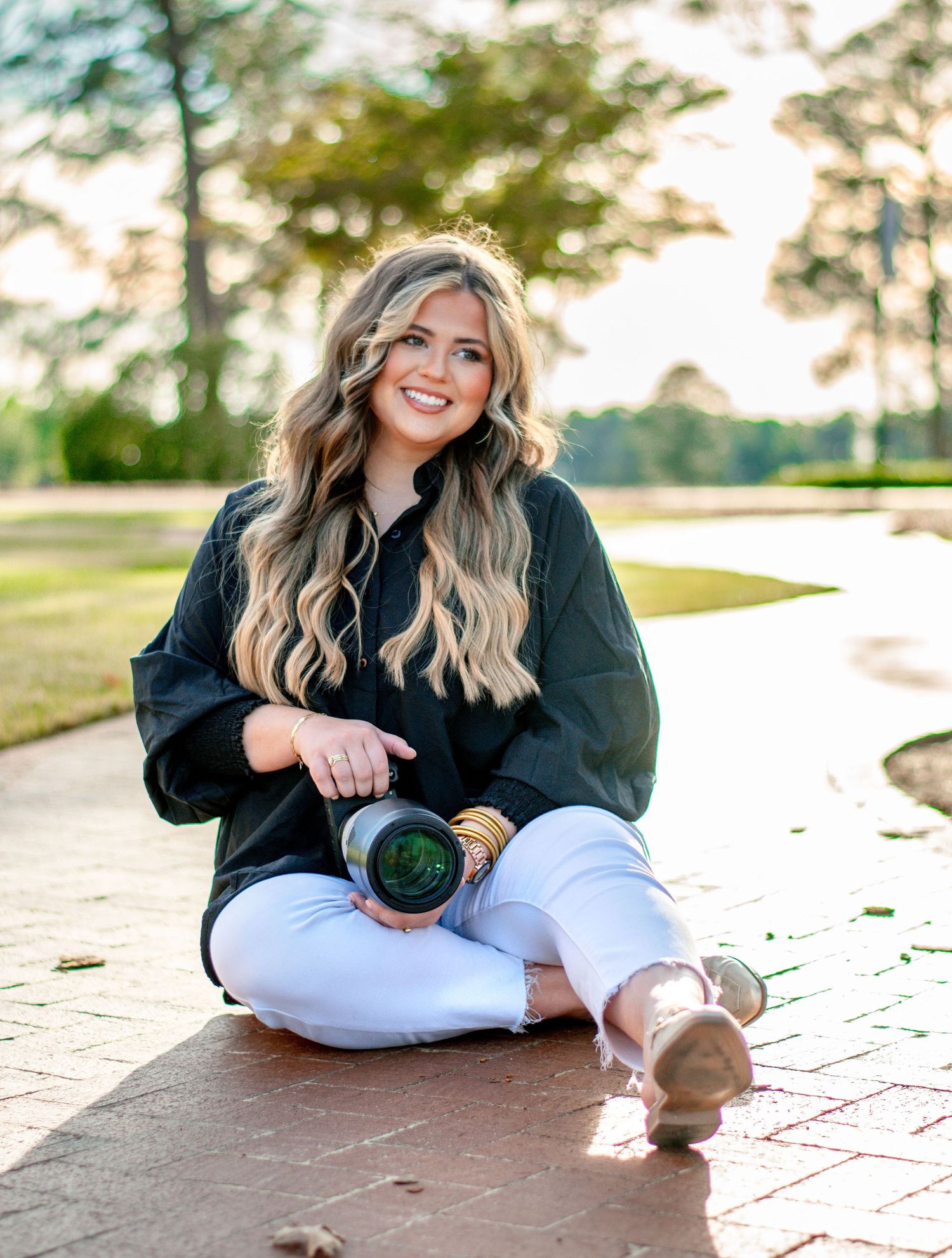 A photographer in casual attire sits cross-legged outdoors with professional camera equipment.