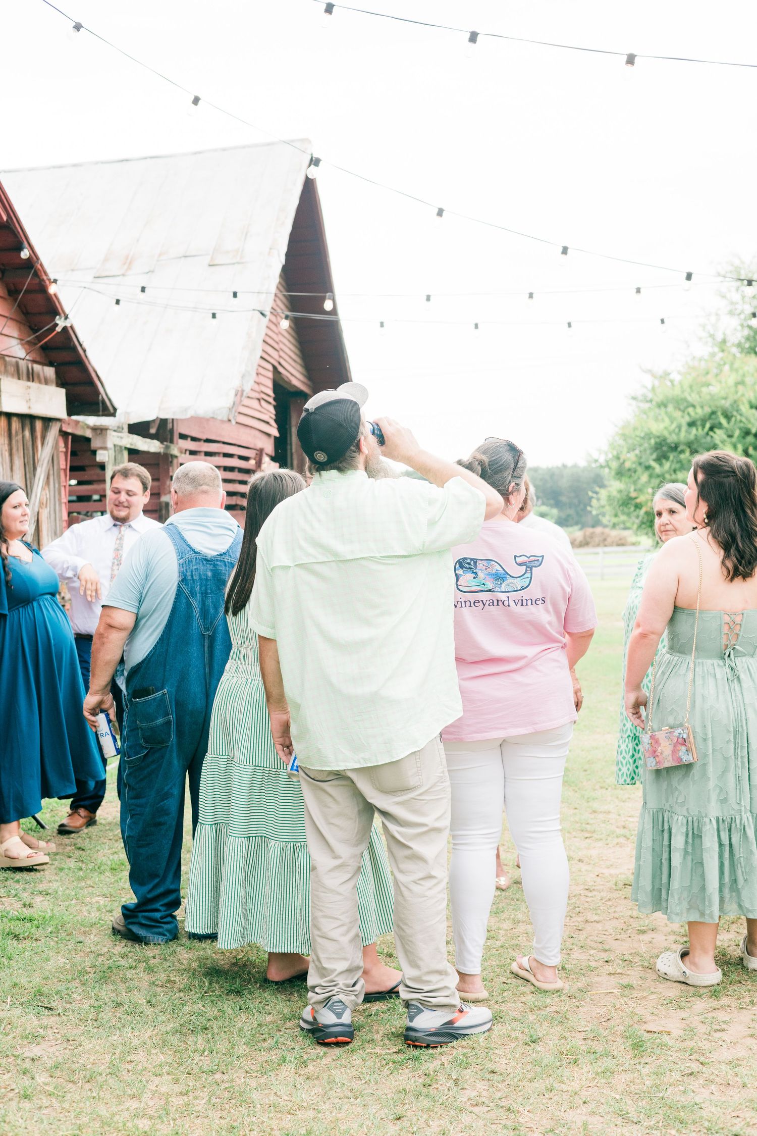 Wedding guests mingling outdoors near a rustic barn with string lights on a warm sunny day.