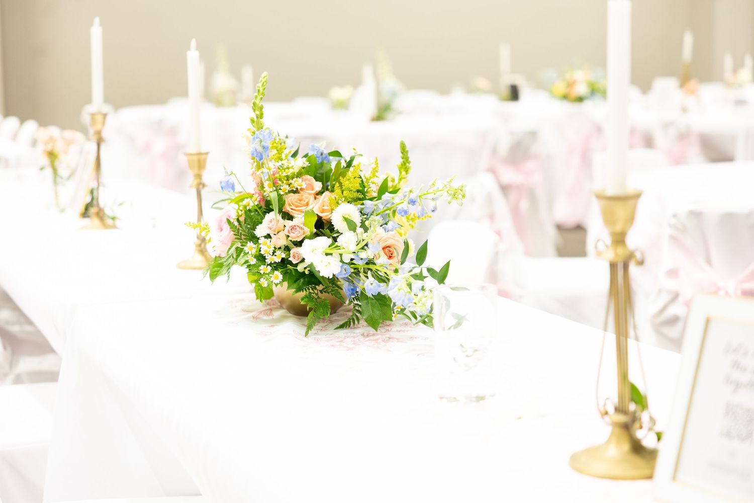 Elegant wedding reception table with white linens, gold candelabras, and a pastel floral centerpiece arrangement.