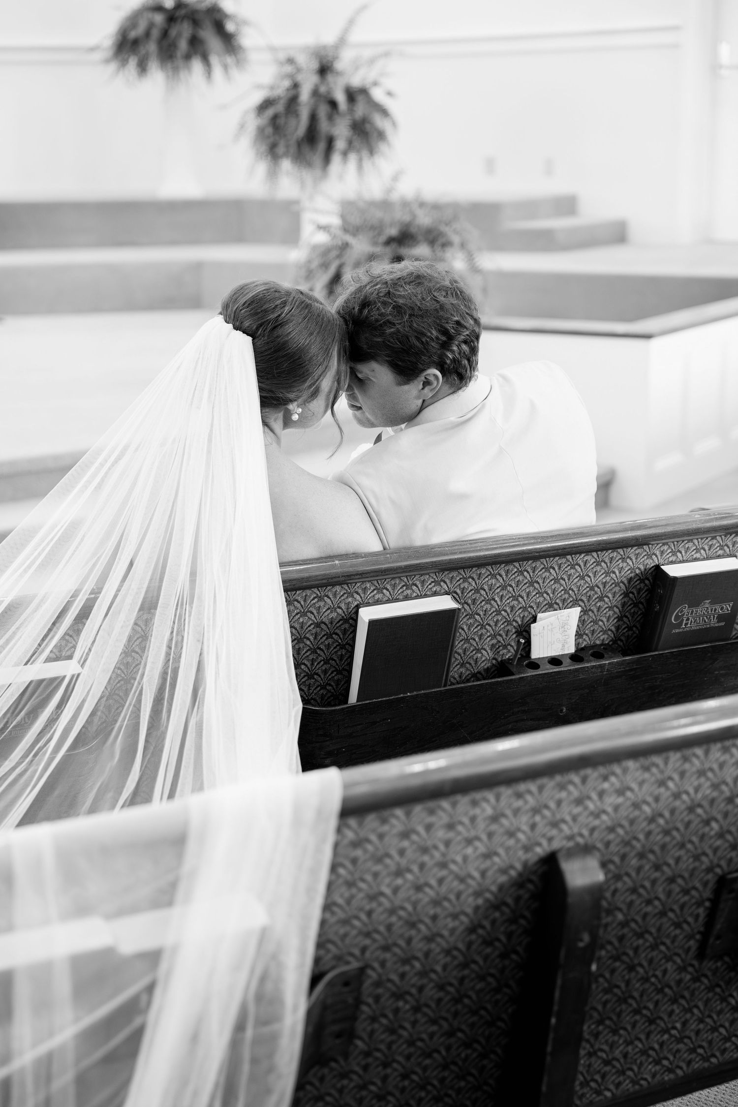 Black and white photo of bride and groom sharing a kiss in a church pew with wedding veil draped over bench.