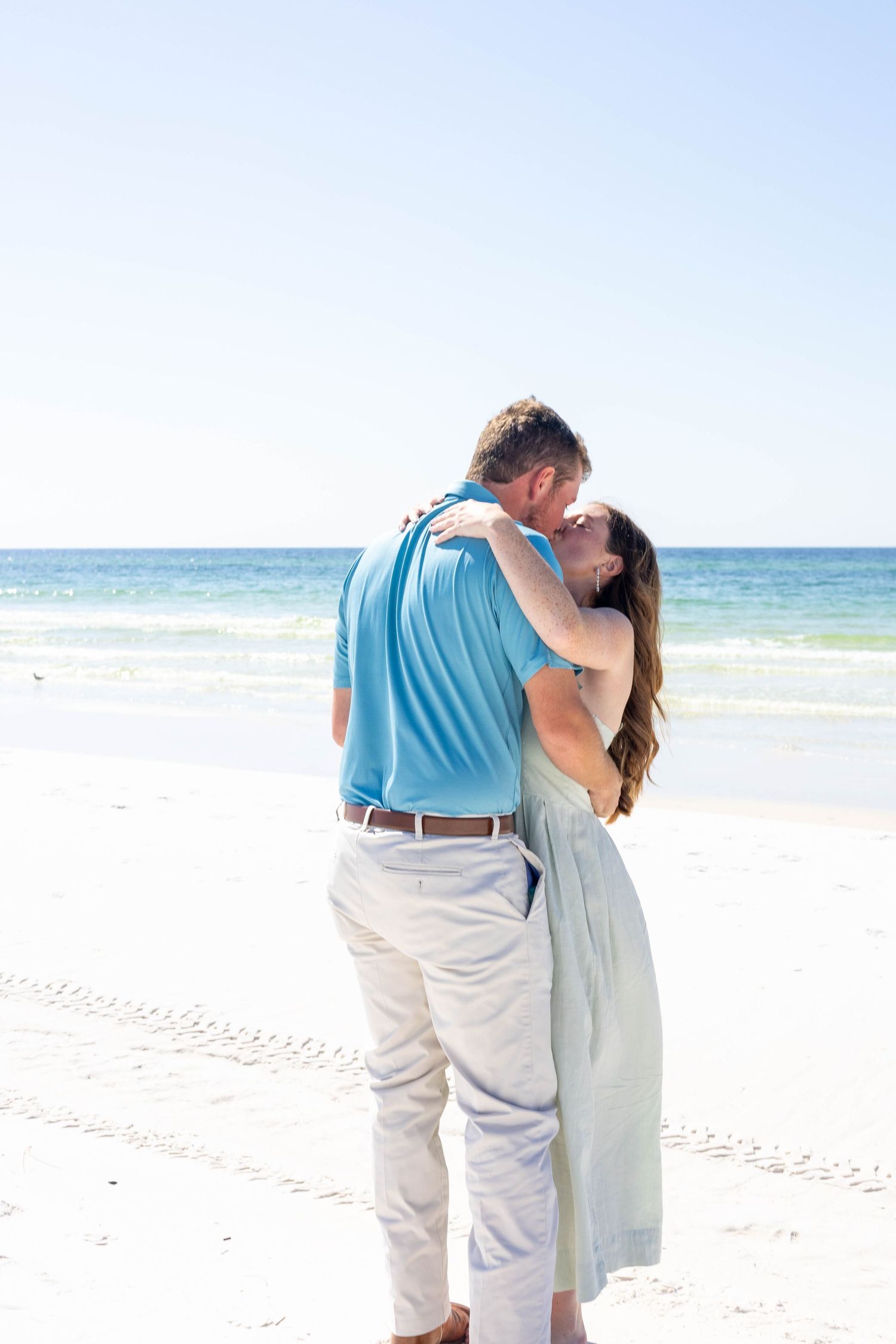 Romantic couple sharing a kiss on a sunny beach with turquoise ocean waves in the background.
