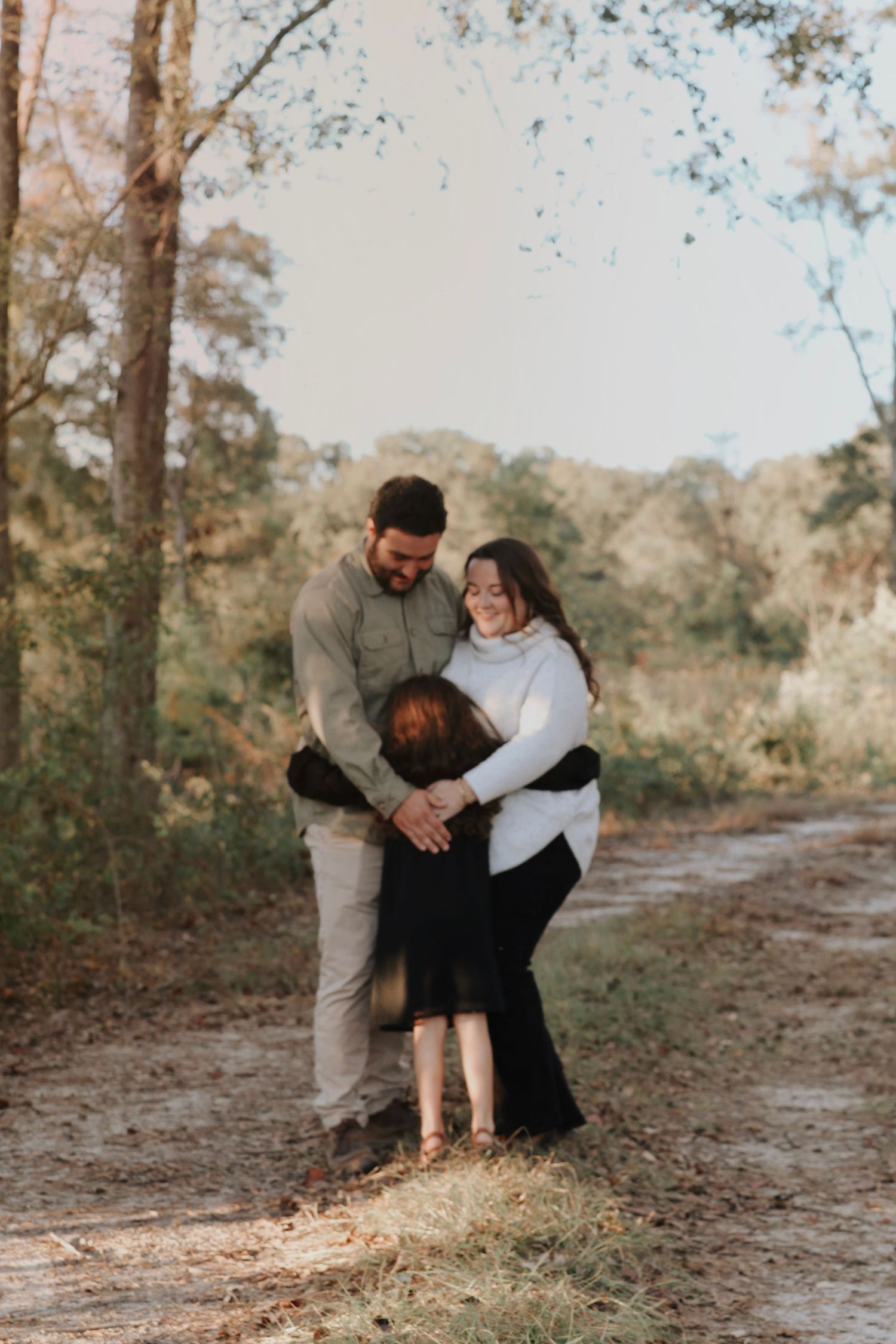 A family of three shares a warm embrace on a sunlit dirt path surrounded by tall trees in a serene outdoor setting.