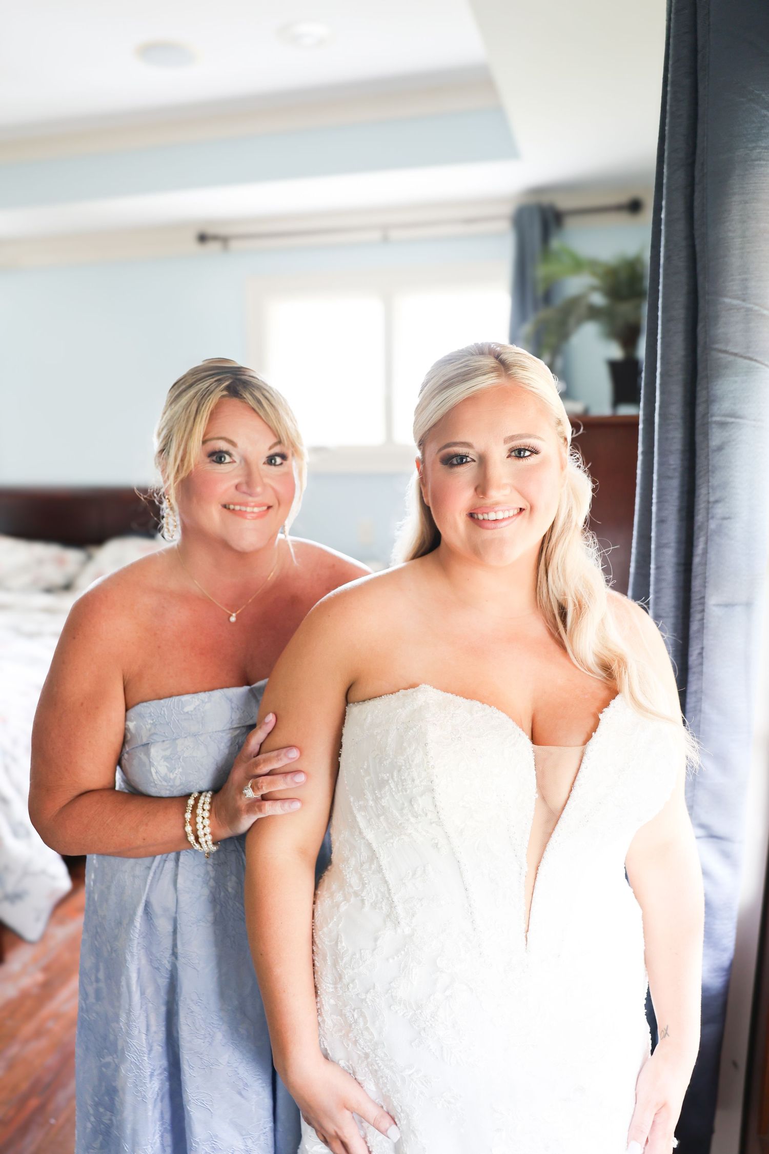 Bride in white lace gown with bridesmaid in blue dress helping her get ready in a bright bedroom.