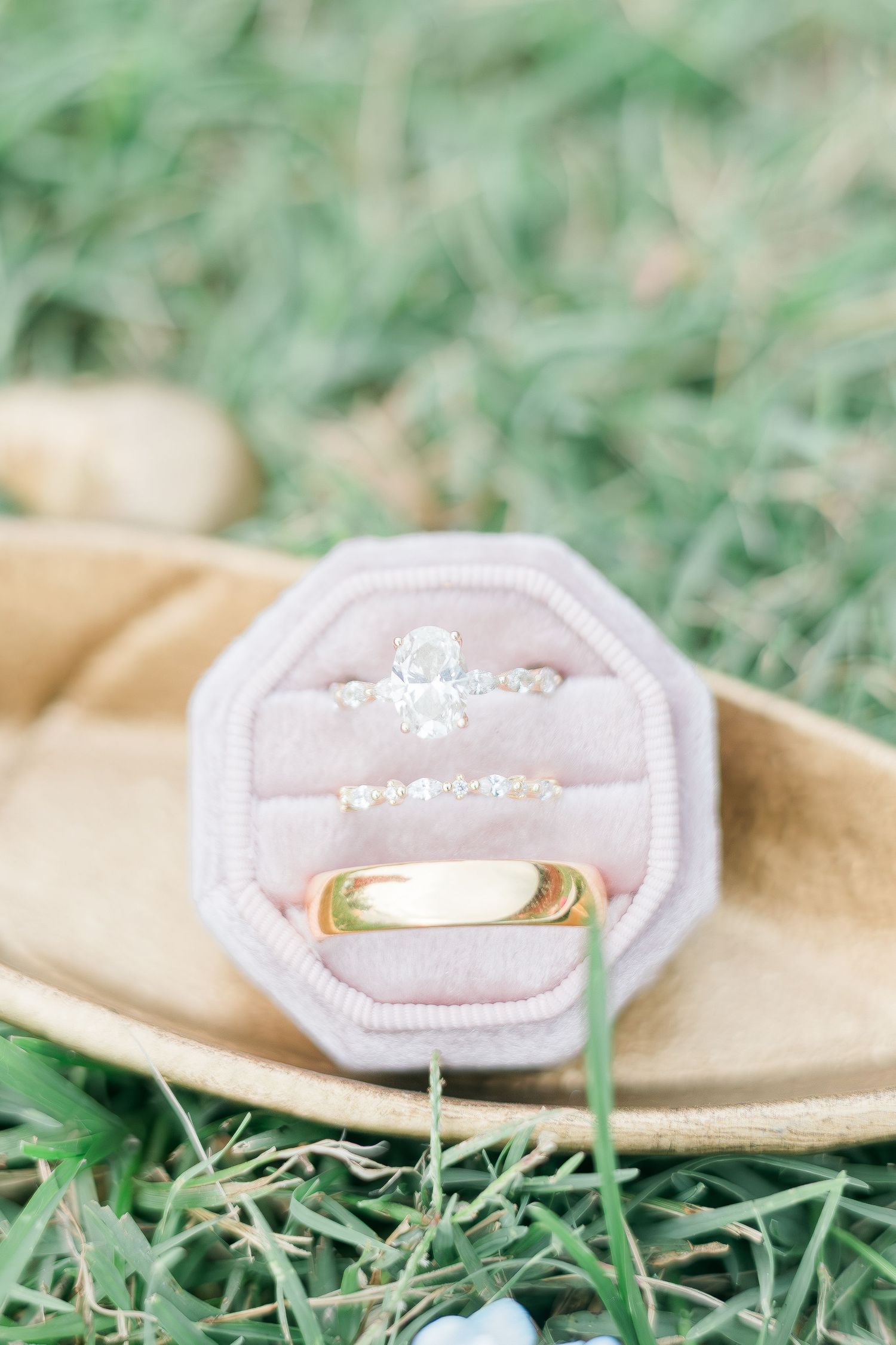 Gold wedding band and diamond engagement ring displayed in a blush pink velvet octagon ring box on grass.