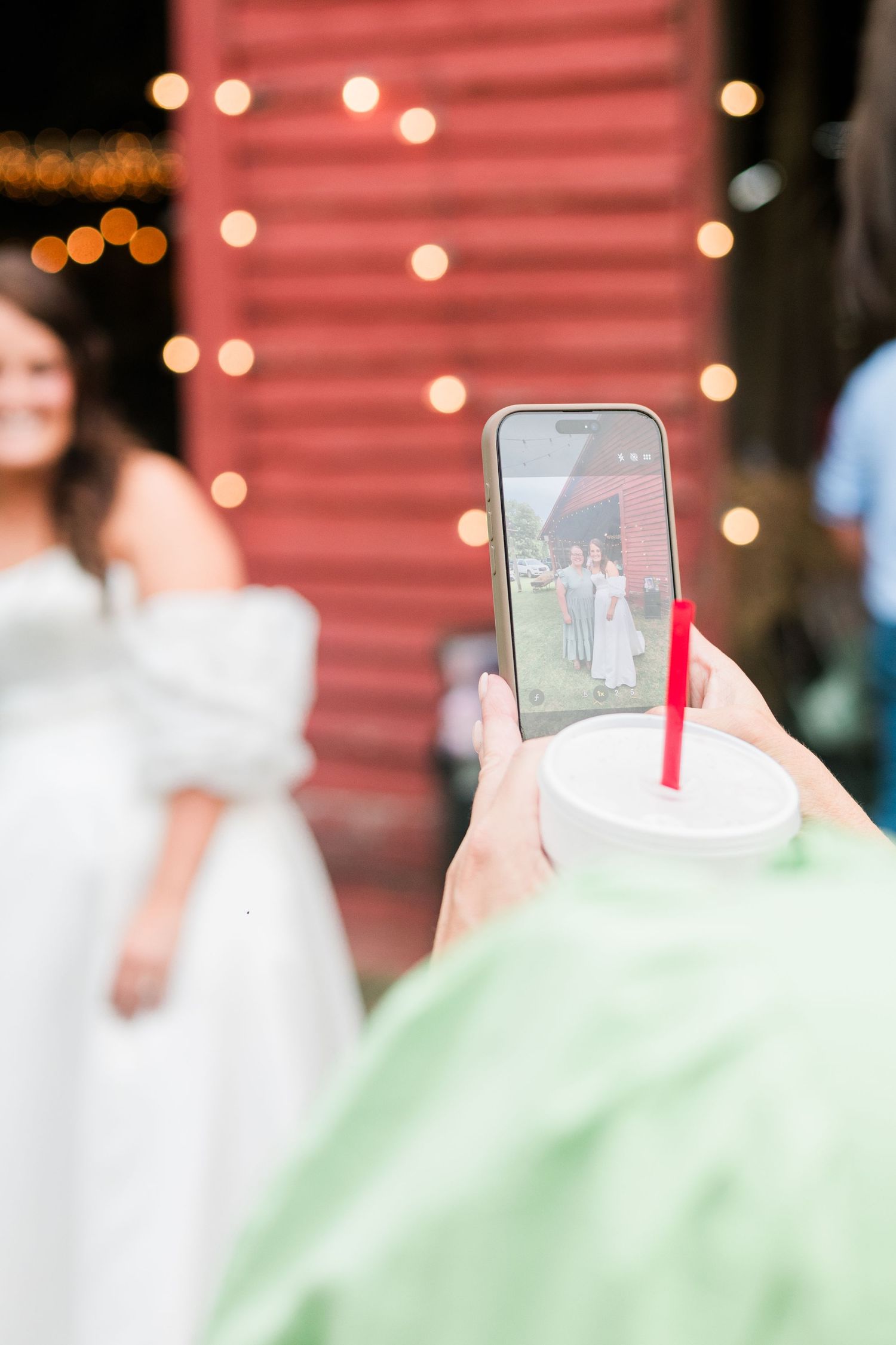 Wedding guest captures bride and wedding party on smartphone near a red barn with string lights.