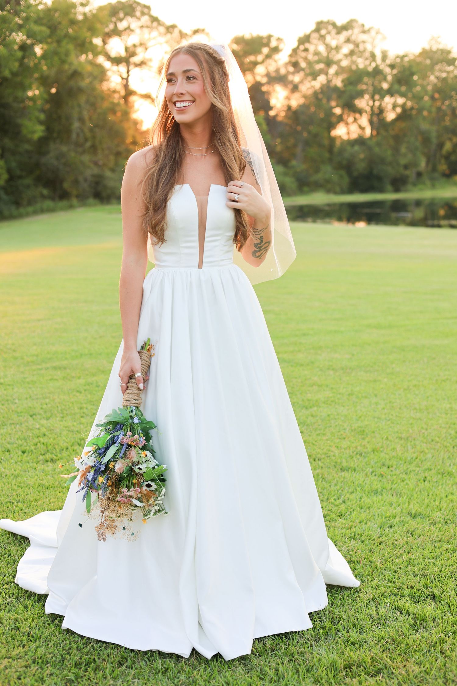 Bride in elegant white gown poses with natural bouquet against green lawn at sunset.