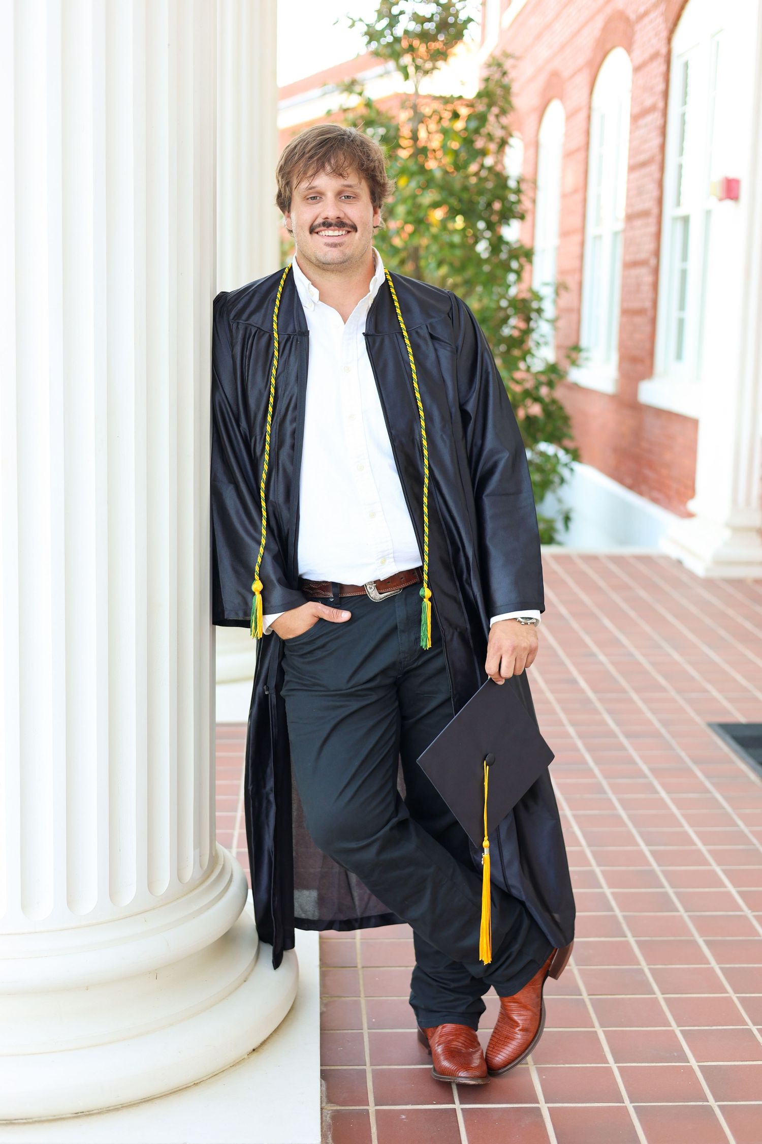 Graduate in black gown with gold cord sitting between white columns.