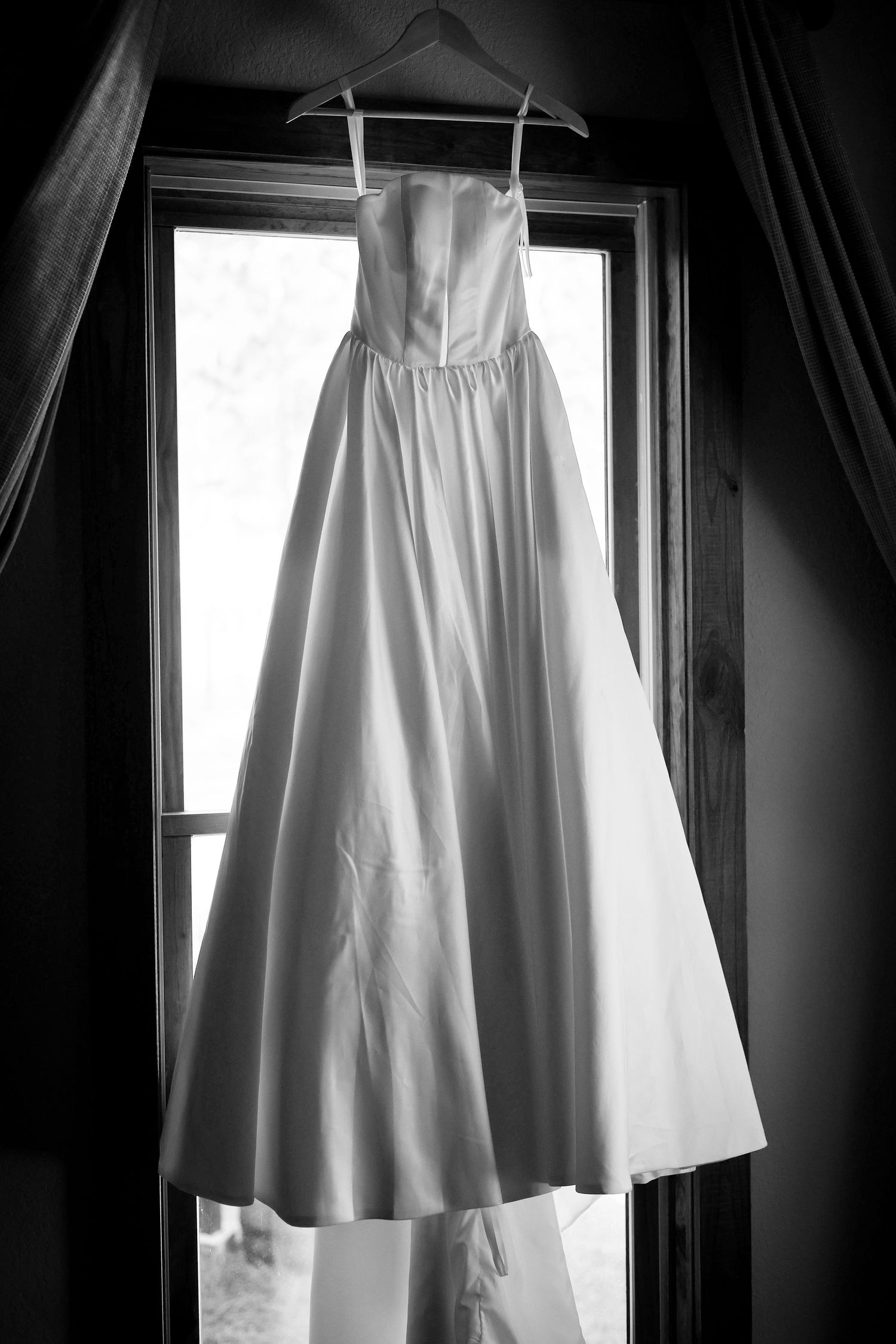 A white wedding dress hangs elegantly against a window in dramatic black and white lighting.