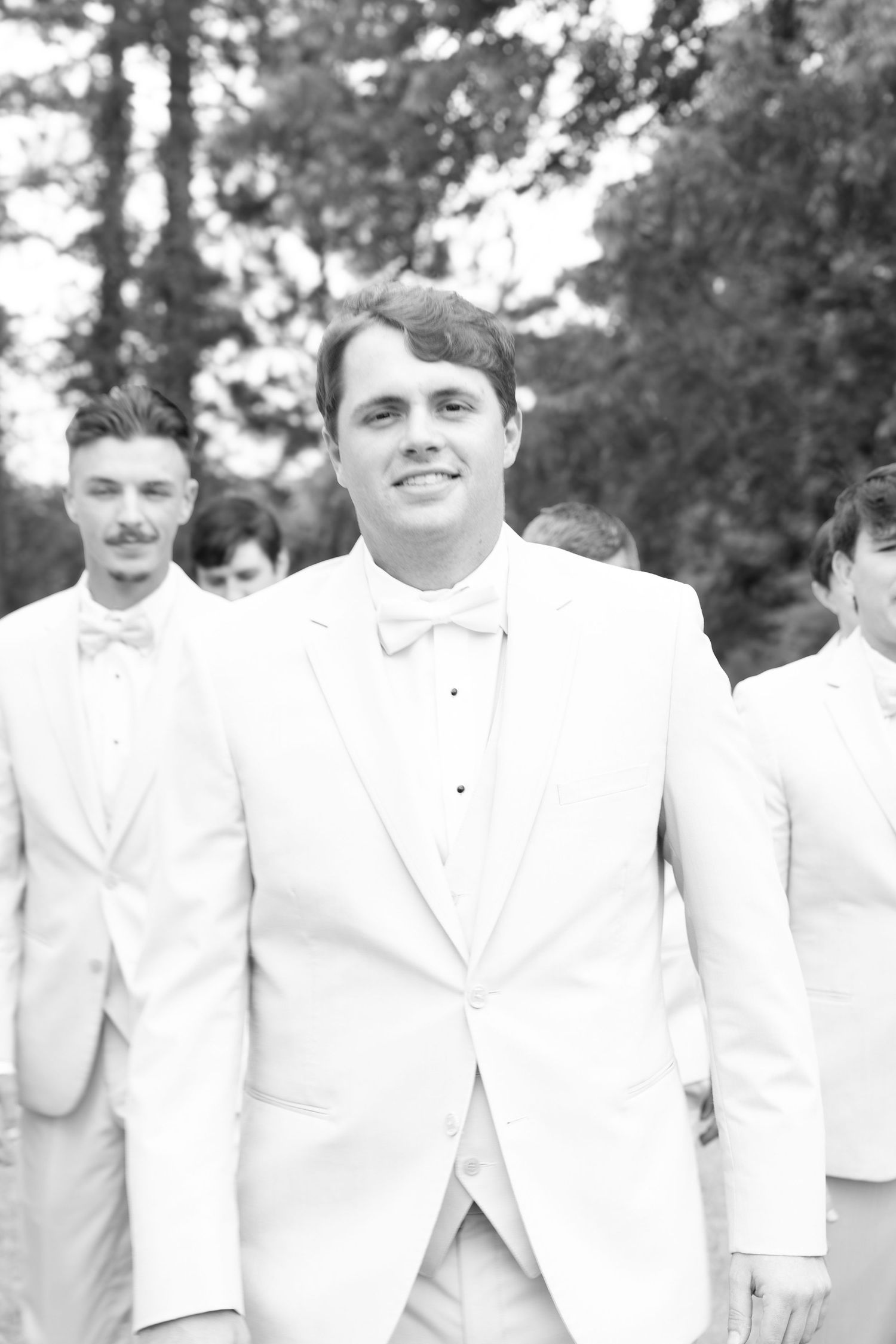Smiling groom in white tuxedo leads groomsmen in black and white wedding photo outdoors.